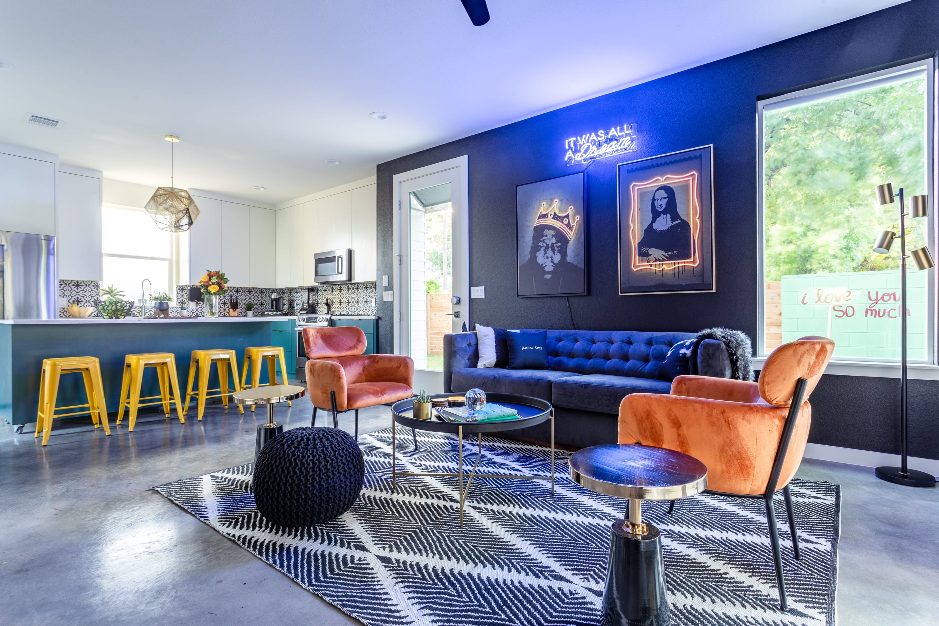 Vibrant living room with a dark blue tufted sofa and two orange velvet armchairs, centered around a black and white geometric rug. A neon sign and pop art adorn the dark accent wall, with a modern kitchen featuring yellow bar stools visible in the background.