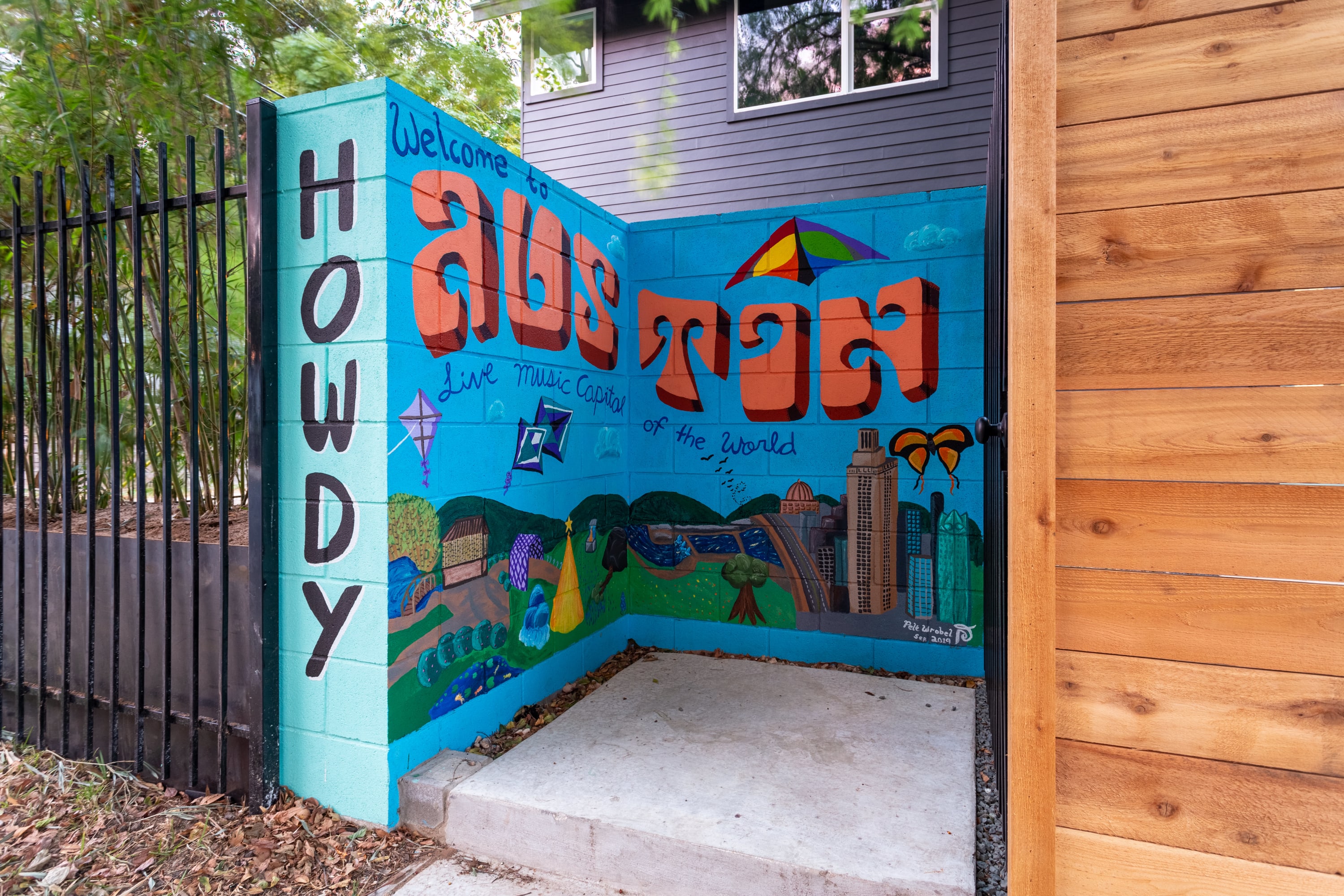 Vibrant exterior mural welcoming guests to Austin, featuring colorful text "Howdy Austin" and "Live Music Capital of the World" alongside a stylized cityscape and natural elements. The mural is painted on a concrete wall next to a black metal fence.