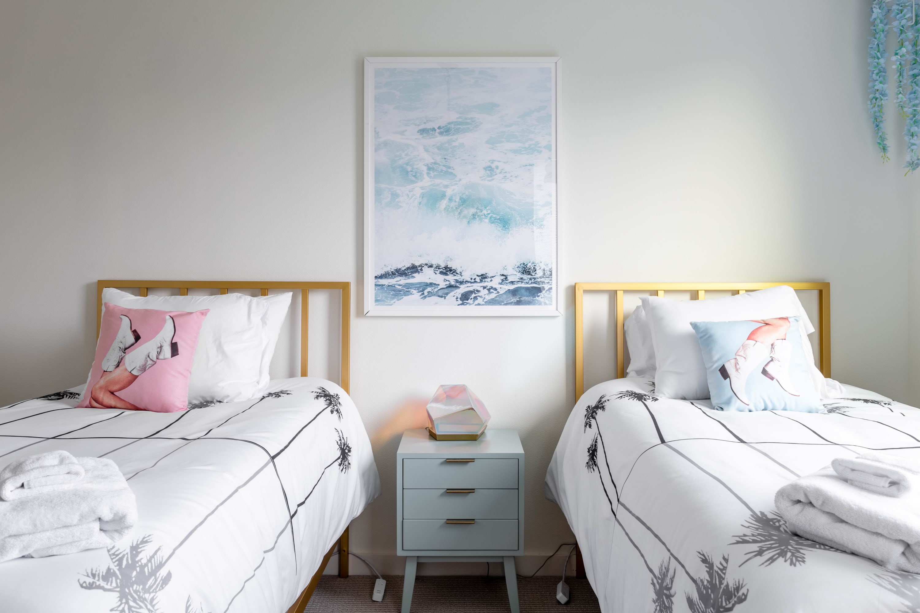 A close-up view of a bright bedroom with two gold-framed twin beds, white bedding with palm tree patterns, and decorative pillows. A light blue nightstand with three drawers and a geometric lamp sits between the beds, beneath a large ocean wave art print.
