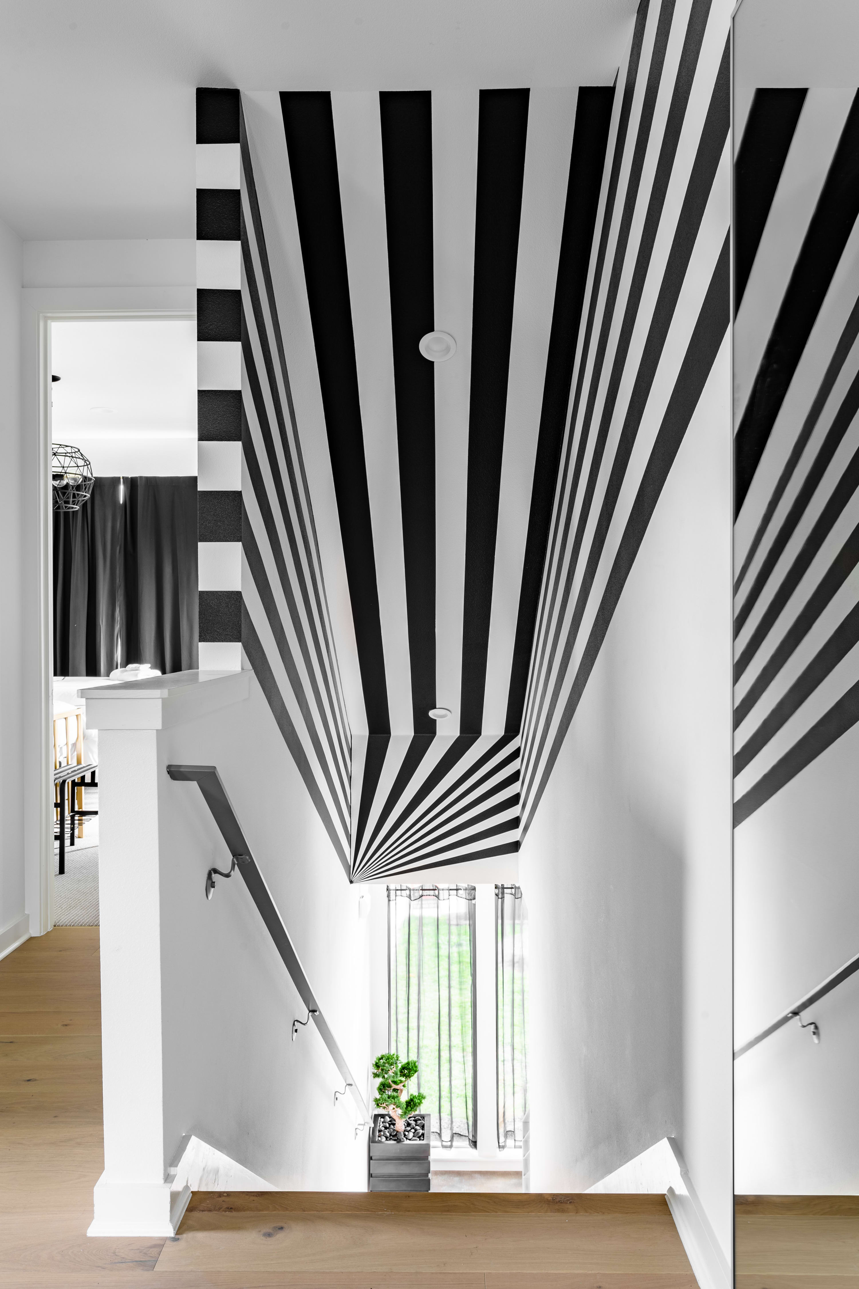A striking staircase features a bold black and white striped ceiling and wall design that creates an optical illusion, leading down to a window with light green curtains and a small potted plant.