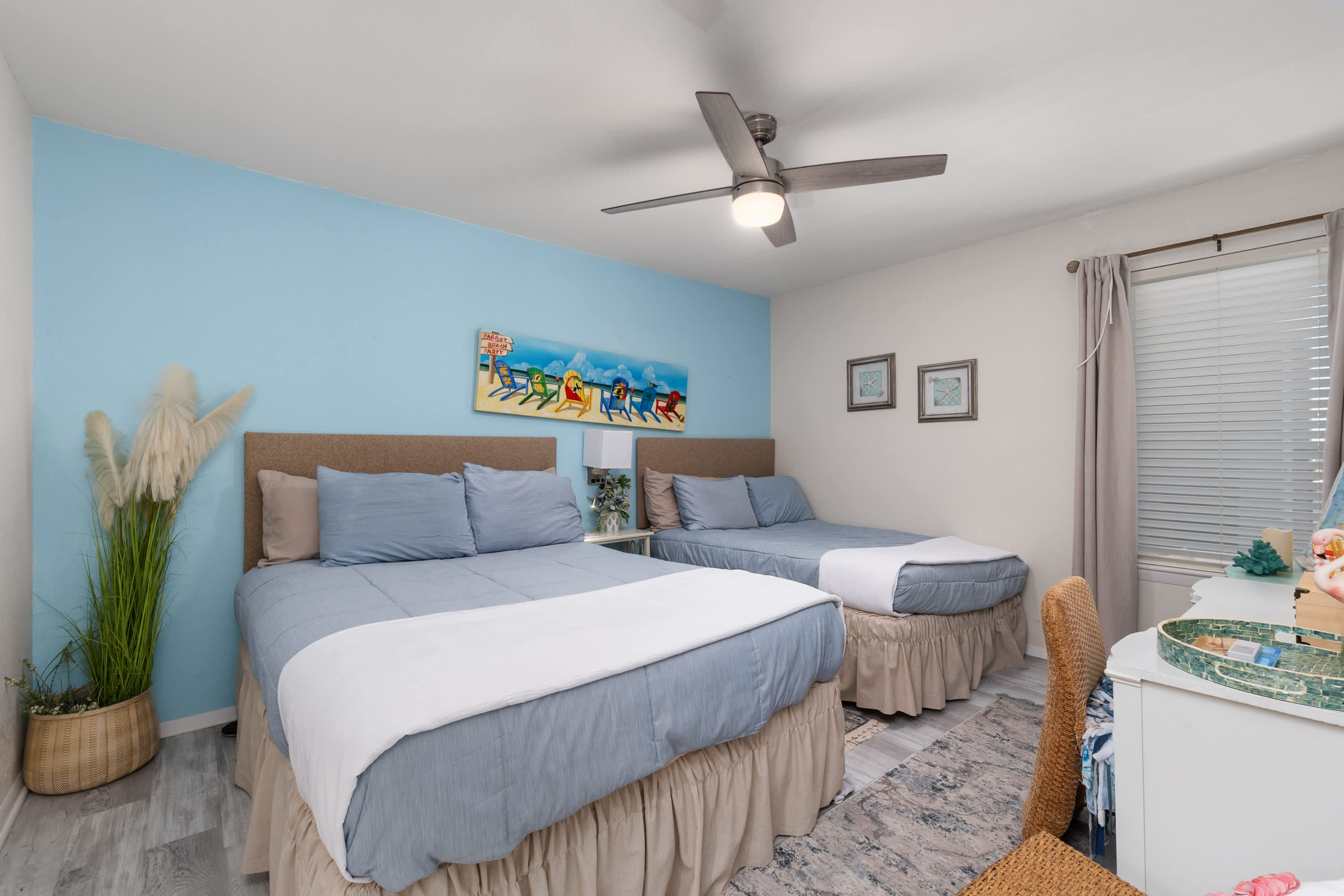 A bright bedroom with a light blue accent wall, featuring a queen bed and a smaller bed, both with blue bedding and light brown bed skirts, under a ceiling fan, with a colorful beach-themed painting above the larger bed.
