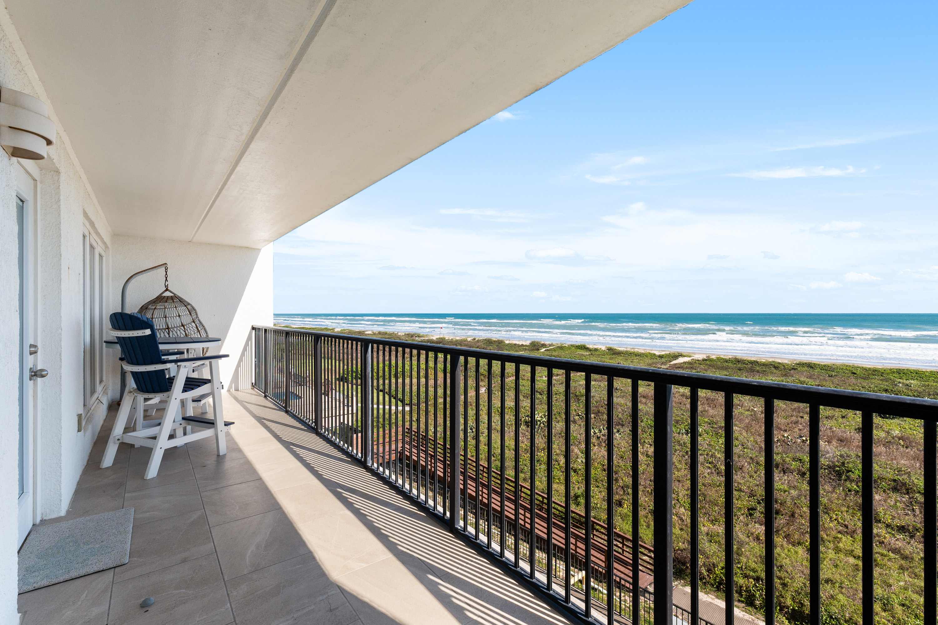 A spacious private balcony with black railings, offering panoramic views of the ocean, sandy beach, and coastal vegetation. White outdoor furniture, including a hanging chair, provides seating.