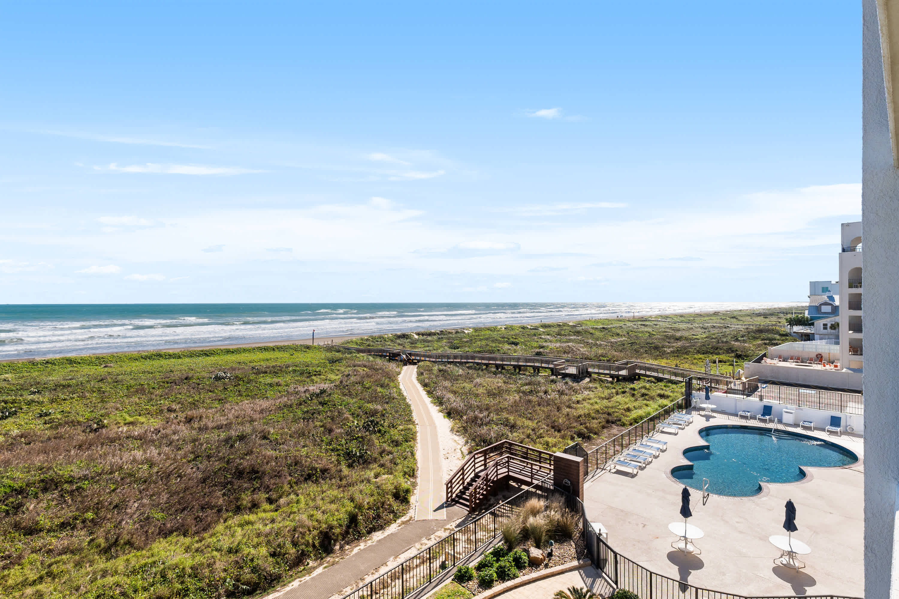 An aerial view showcasing a large outdoor swimming pool with lounge chairs, a winding boardwalk leading to a sandy beach, and the vast ocean under a clear blue sky. Lush green vegetation separates the property from the beach.