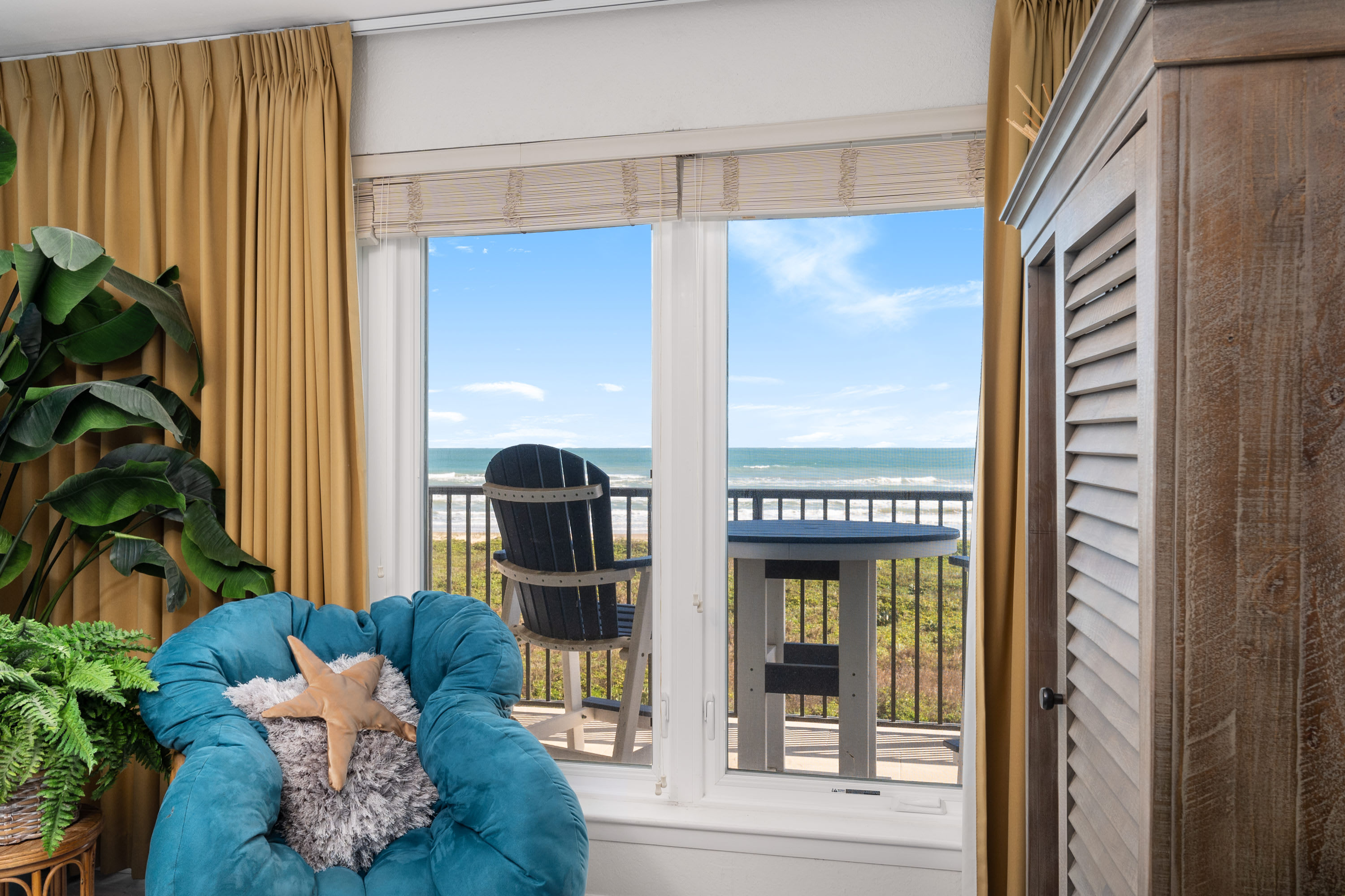 A cozy corner in a bedroom featuring a teal bean bag chair with a starfish pillow, surrounded by potted plants. A large window offers a clear view of the ocean and a balcony with outdoor seating.