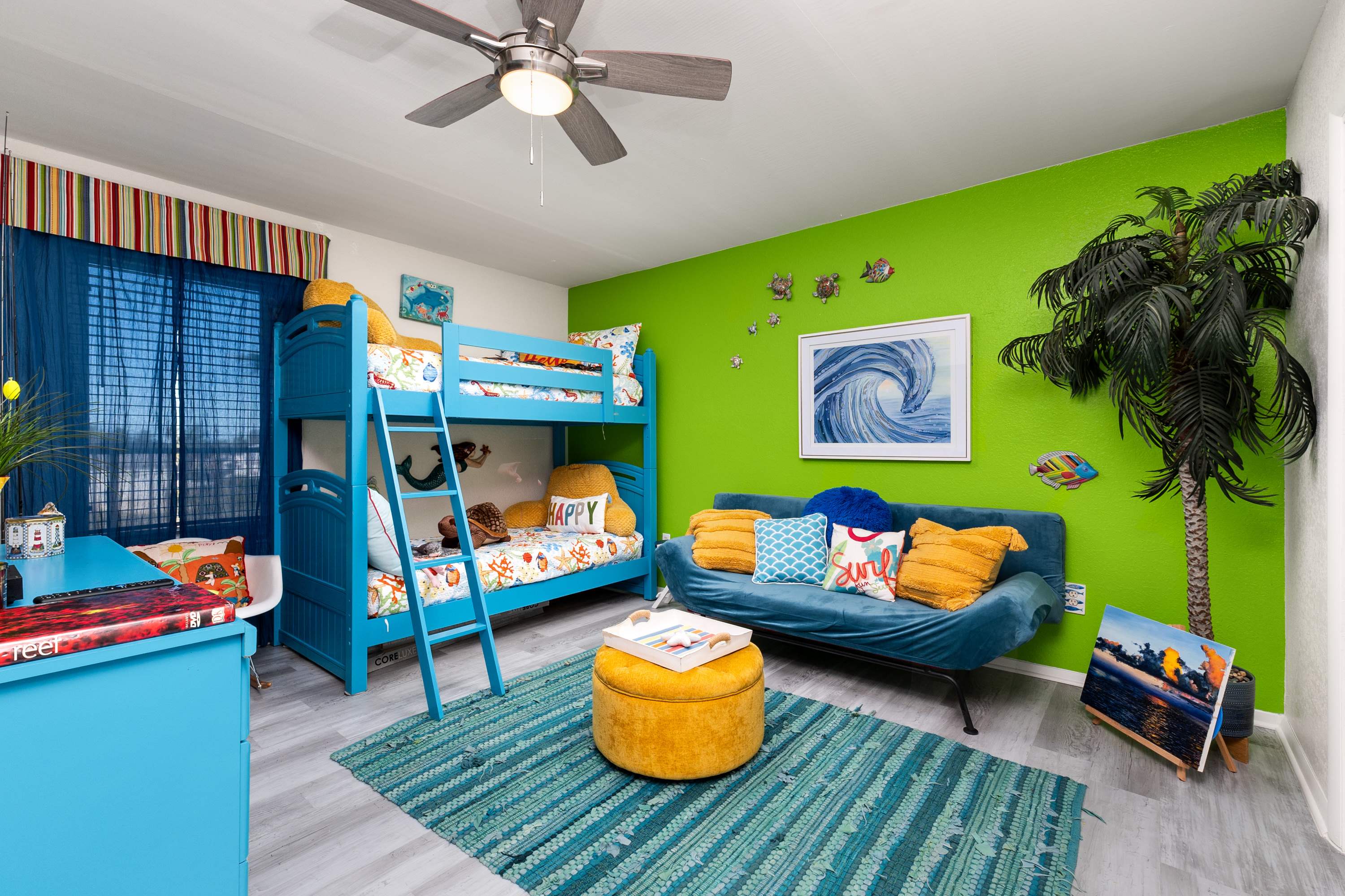 A lively kids' bedroom with a bright green accent wall, featuring a blue bunk bed, a blue sofa bed with yellow pillows, a colorful striped rug, and a large wave artwork, creating a playful, ocean-themed space.