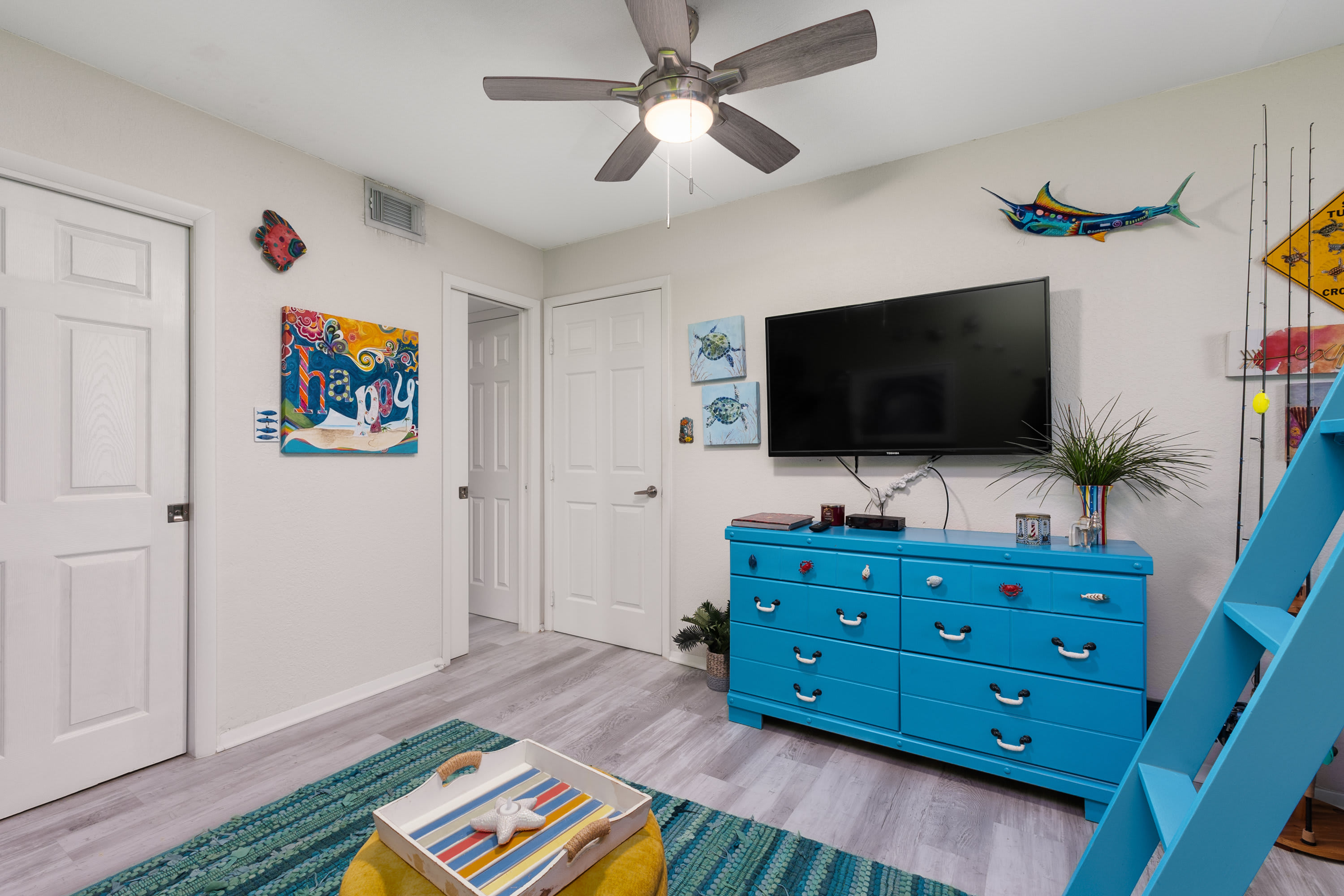 A bright kids' bedroom featuring a large blue dresser with a flat-screen TV mounted above it, colorful fish and fishing-themed wall decor, and a blue ladder leading to a bunk bed (partially visible), all on light wood-look flooring.