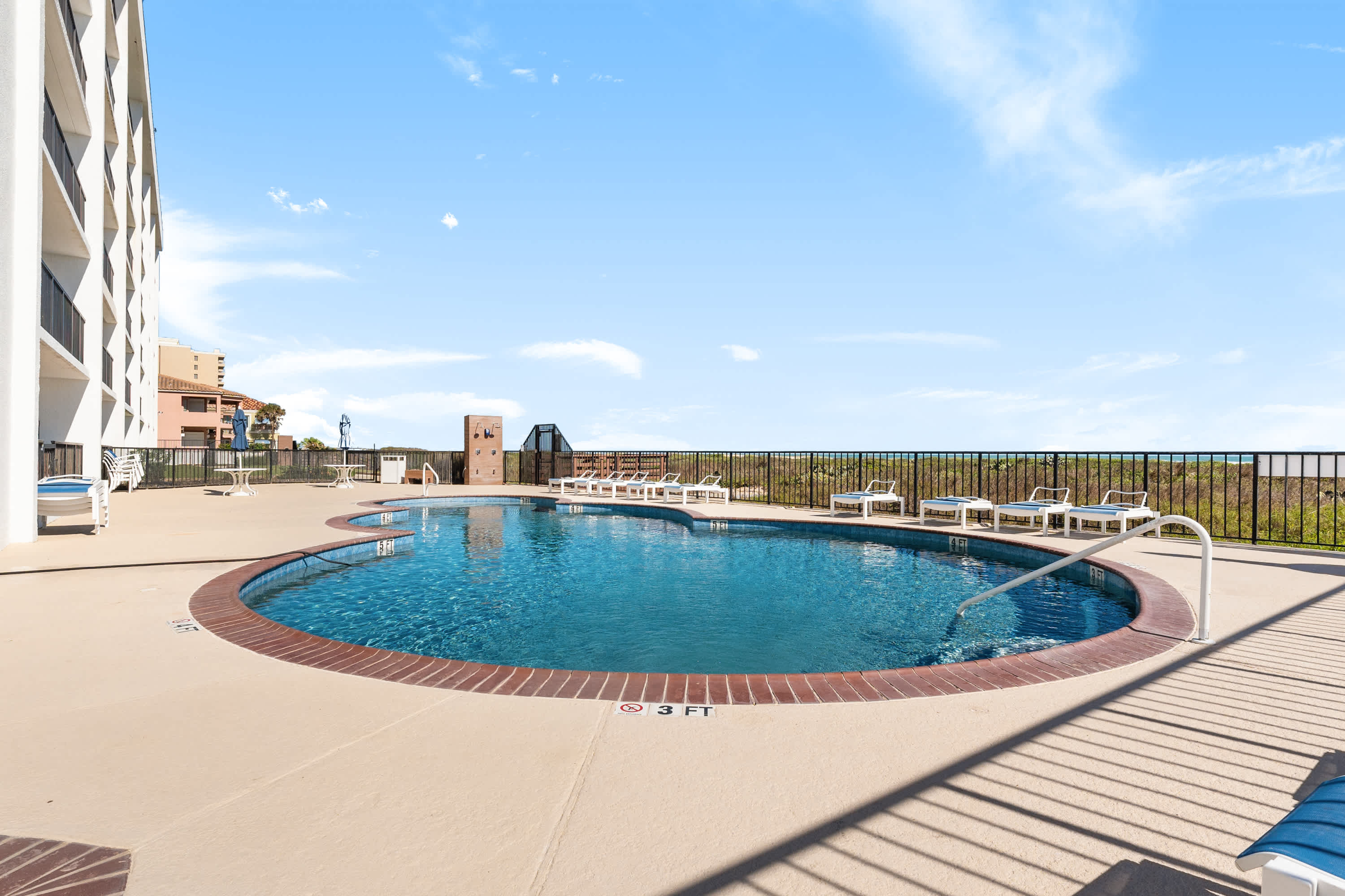 An inviting outdoor swimming pool with a unique clover-like shape, surrounded by lounge chairs on a concrete deck, with a clear blue sky overhead and glimpses of the ocean in the distance.