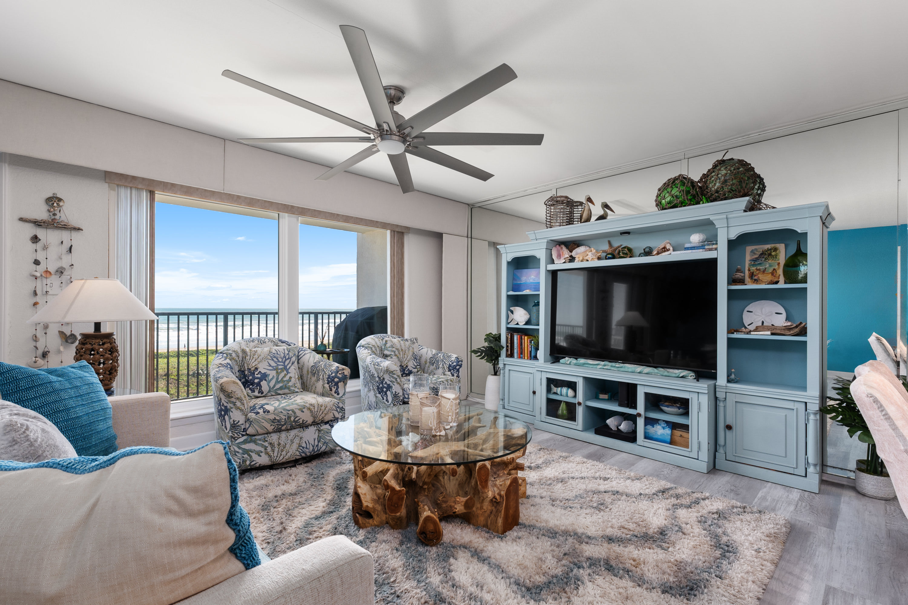 A spacious living room with a large light blue entertainment center housing a flat-screen TV, two floral armchairs, and a glass-top coffee table with a unique wooden base. Mirrored walls reflect the space and ocean view.