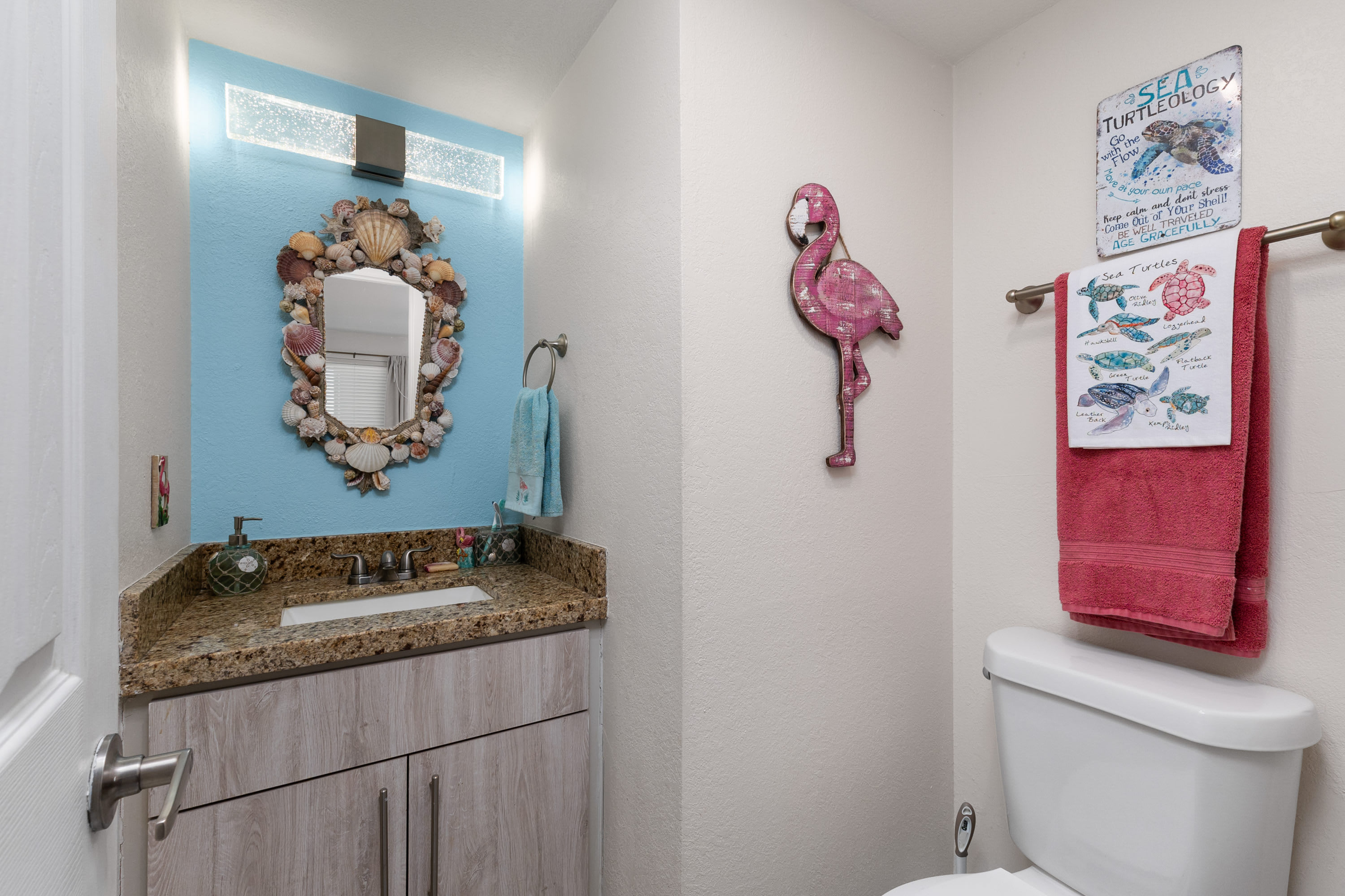 A small, brightly lit bathroom with a light blue accent wall behind a single vanity, featuring a shell-encrusted mirror, a toilet, and a pink flamingo wall decoration, creating a whimsical, beach-themed powder room.