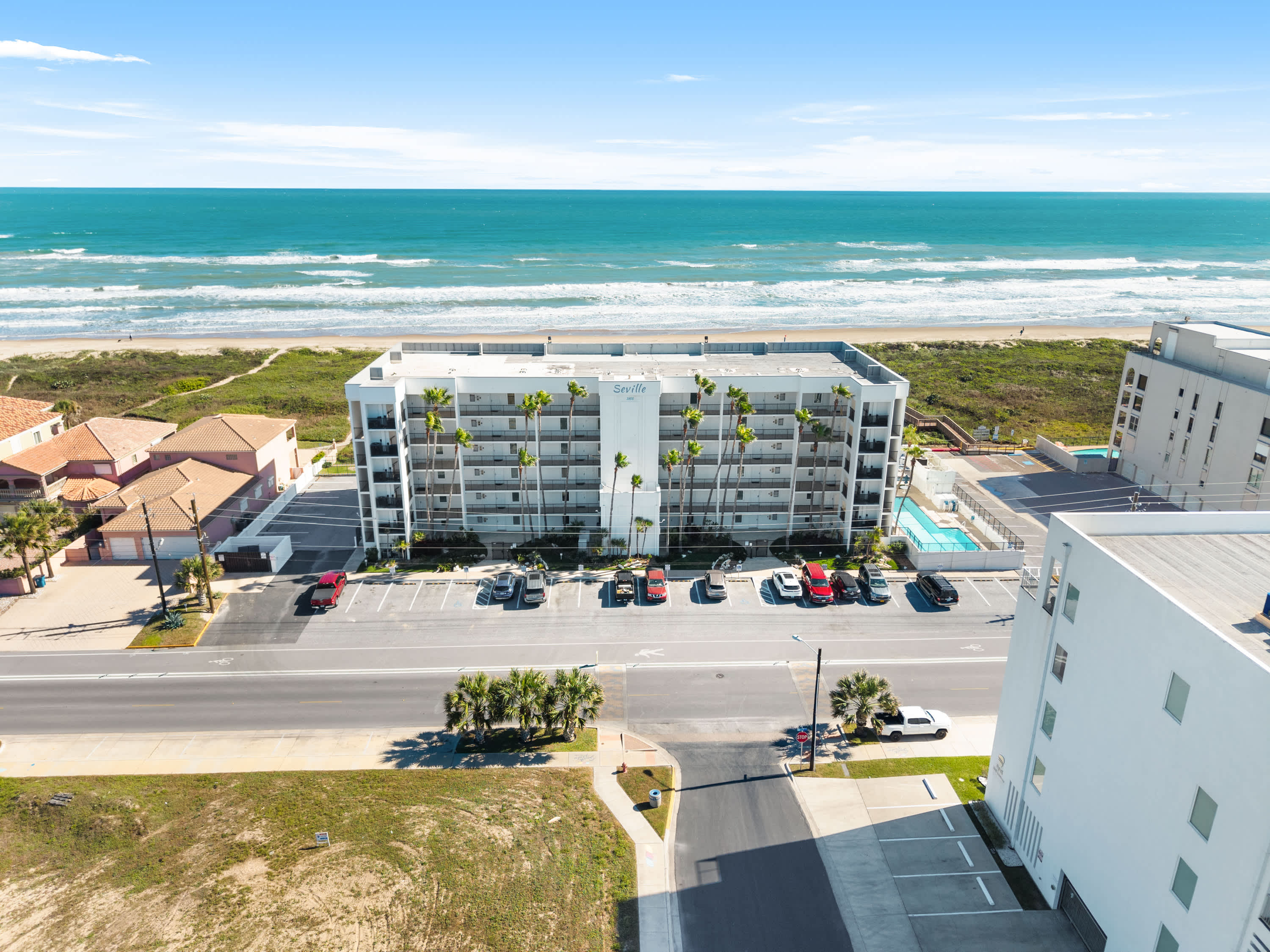 A wide aerial shot of a white beachfront condominium building with palm trees, facing the expansive blue ocean with white waves, surrounded by parking and other buildings under a clear sky.