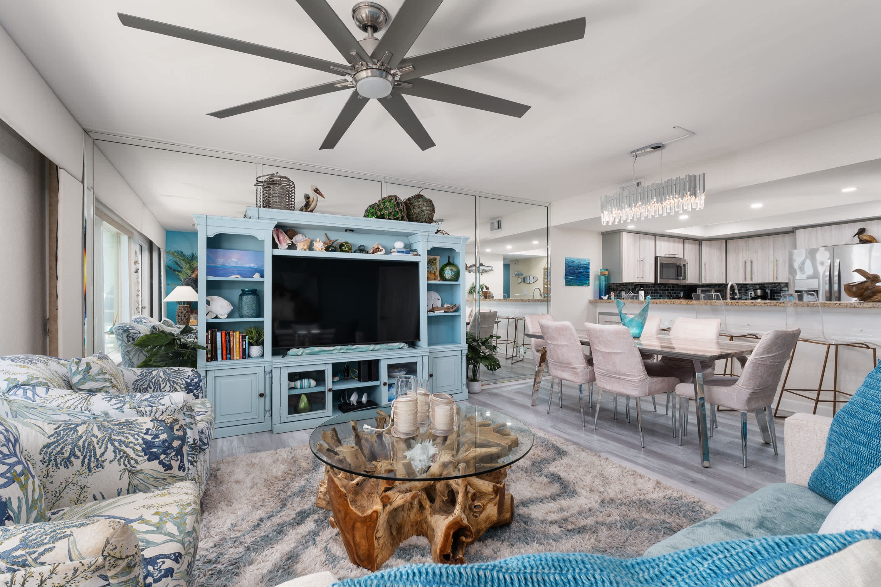 A wide shot of the living room and dining area, featuring a large light blue entertainment center, a glass-top coffee table, and a dining table with pink chairs. Mirrored walls expand the perception of space.