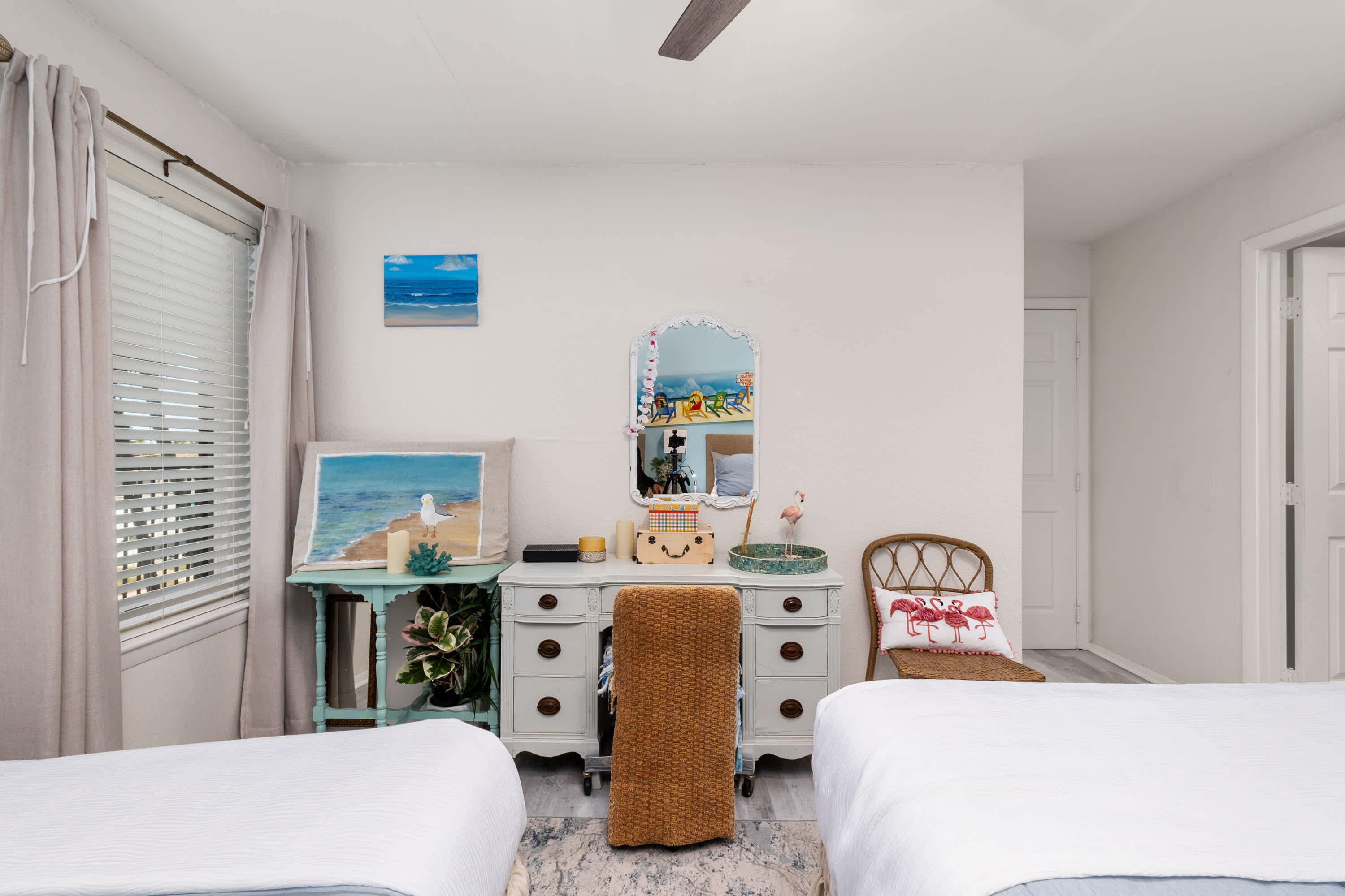 A light-filled bedroom corner featuring a white dressing table with a shell-framed mirror, a wicker chair, and two beds with white bedding, next to a window with blinds and light curtains.
