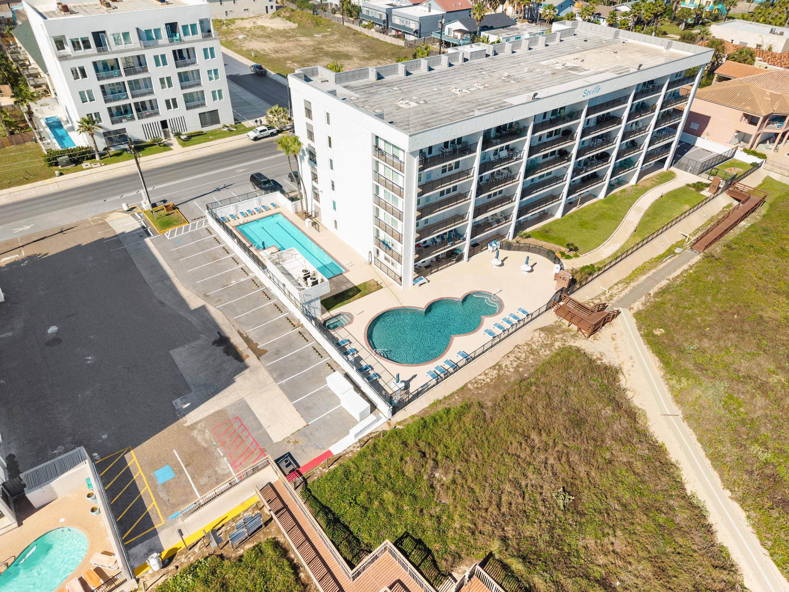 An aerial view showcasing a multi-story white condominium building with multiple pools and a parking lot, situated near a grassy dune area and the ocean, under a bright blue sky.