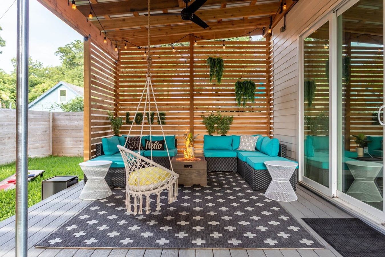 Spacious covered outdoor patio with slatted wooden walls, featuring a turquoise sectional sofa, a fire pit, and a macrame swing chair. A geometric rug covers the deck, and string lights are strung across the ceiling.