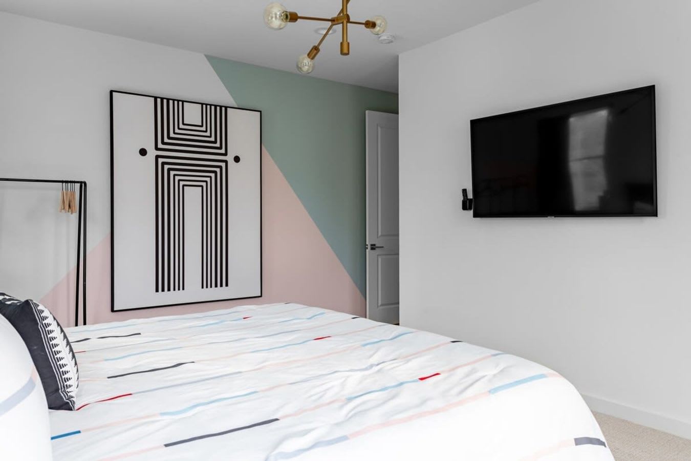 Another view of Bedroom-1, showing a wall with a large abstract black and white artwork and a geometric color-blocked accent wall in pink and mint green. A wall-mounted TV is visible on the right.