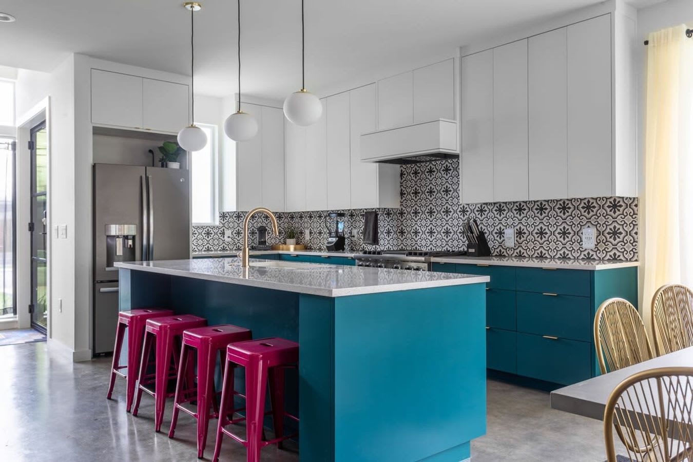 Wide shot of a modern kitchen featuring a teal island with a white countertop and hot pink bar stools. White upper cabinets and teal lower cabinets complement the black and white patterned tile backsplash. Stainless steel appliances are visible.