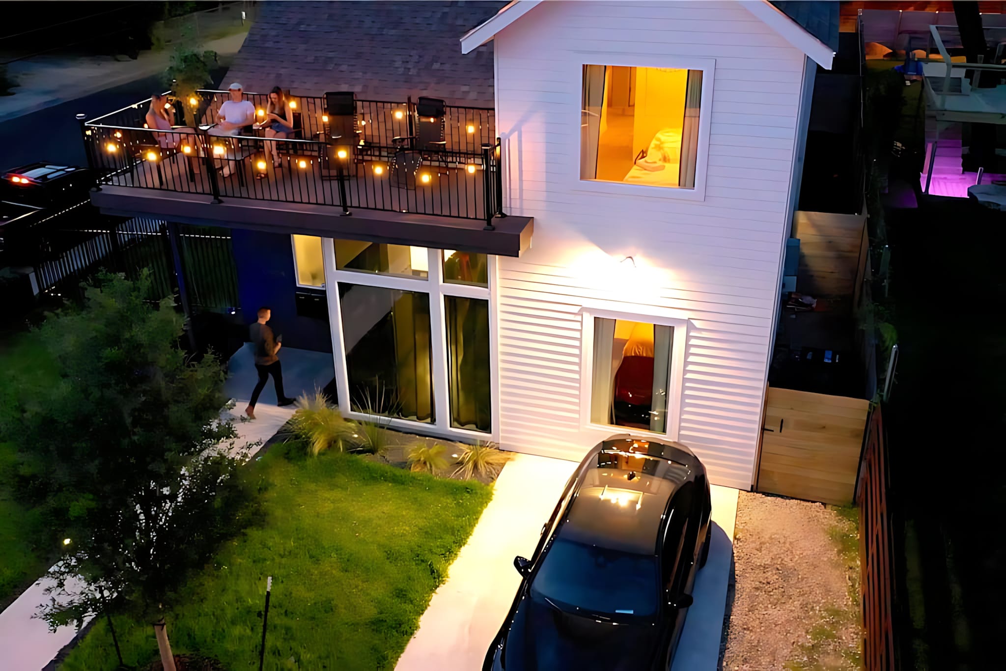 Aerial view of a modern white two-story house with a large second-story balcony where people are gathered. The property features a small lawn, a driveway with a car, and string lights illuminating the balcony at dusk.