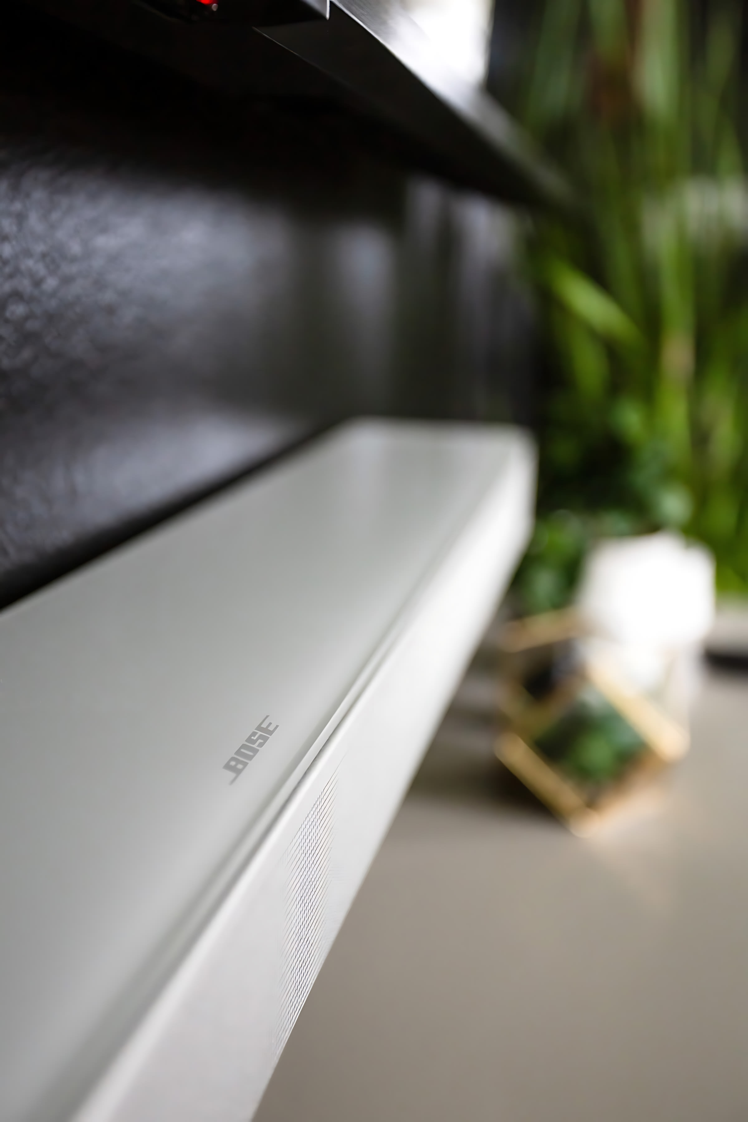 Close-up view of a white Bose soundbar positioned on a white media console, with a blurred background showing a green plant and decorative items. The focus is on the sleek design of the audio equipment.