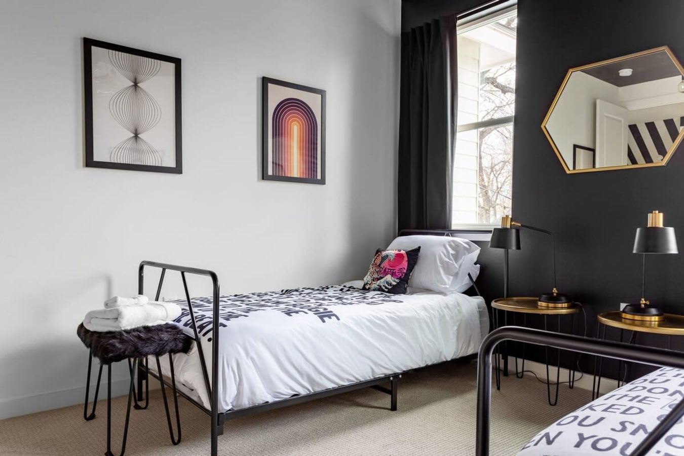 Another perspective of the bedroom with two black metal twin beds, showcasing the white bedding with black text and a colorful abstract art piece on the white wall. A black accent wall with a gold mirror is visible.