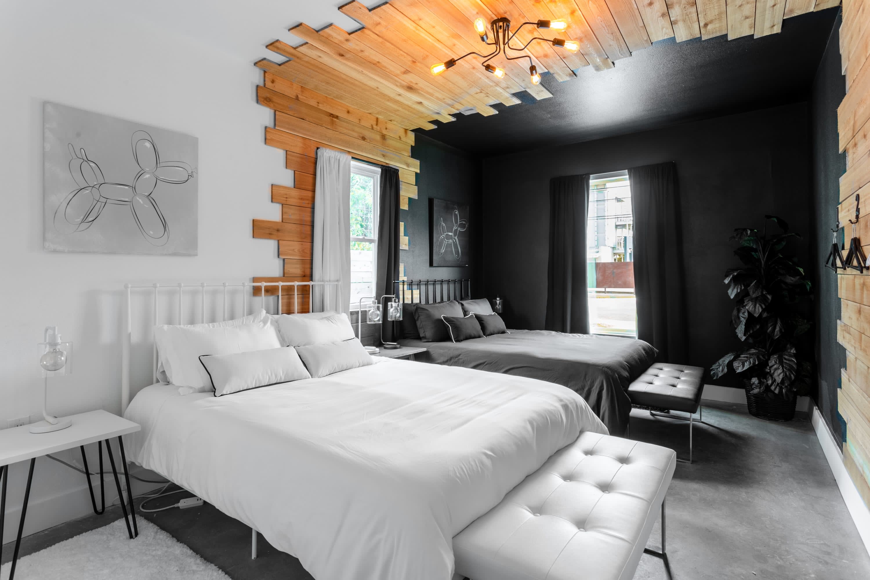 A striking bedroom with two beds, one white and one black, separated by a wooden accent wall and ceiling. The room features a black painted ceiling with a modern light fixture and a large plant in the corner.