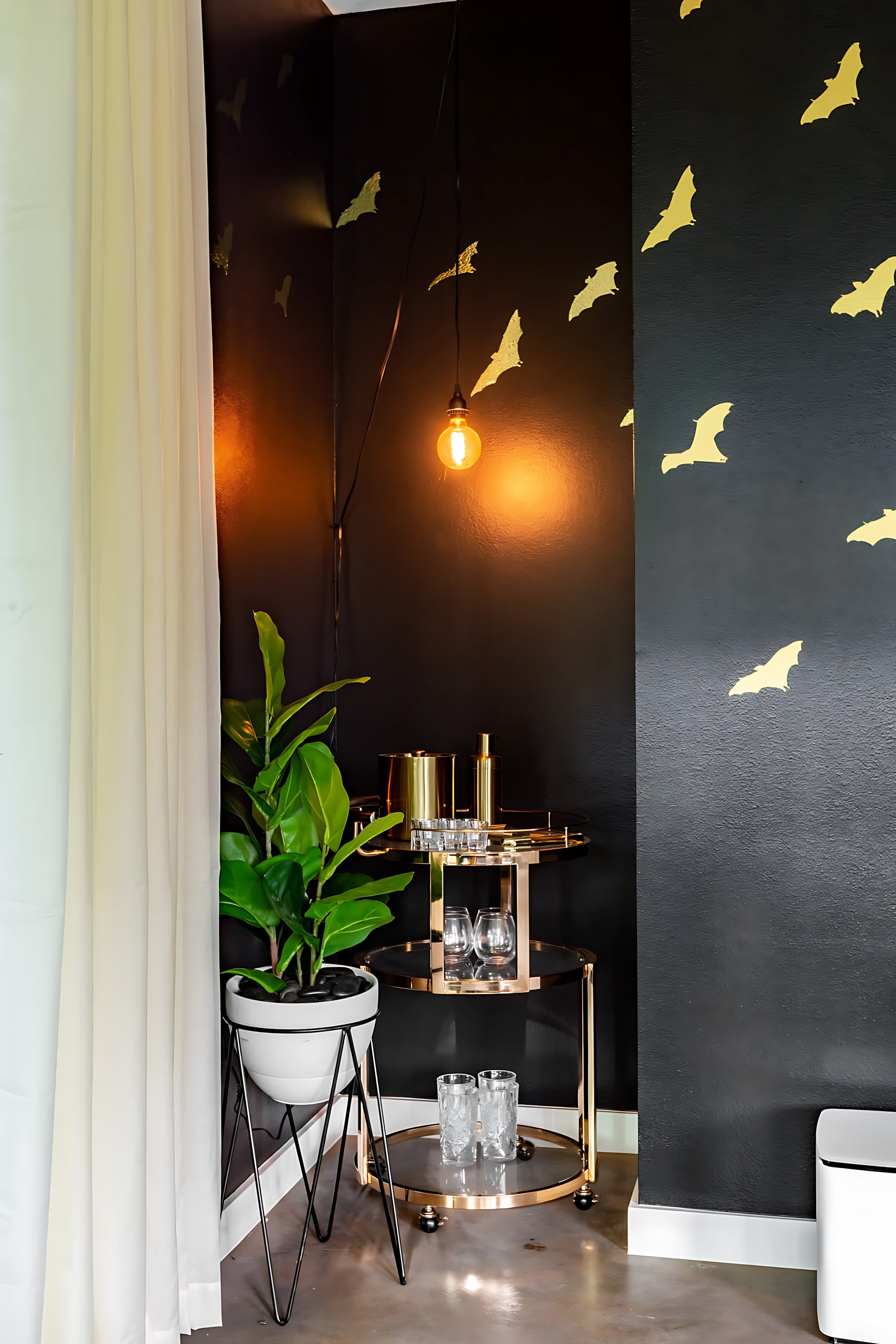 Stylish corner of a living space featuring a gold bar cart stocked with glassware and bottles, next to a large potted plant. The black wall is adorned with golden bat decals, and a single Edison bulb hangs from the ceiling.