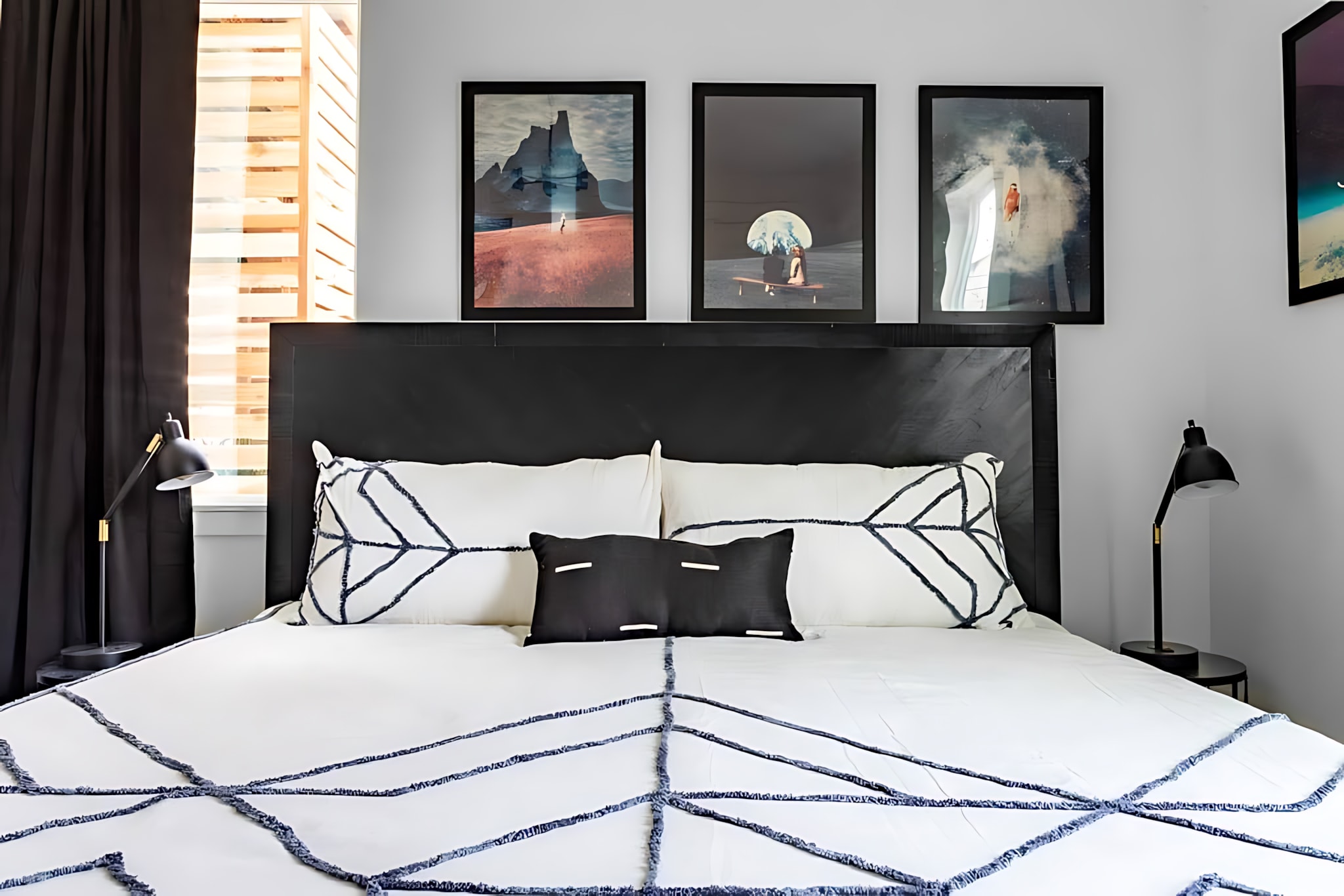 A modern bedroom featuring a black headboard, white bedding with black geometric patterns, and three framed abstract art pieces above the bed. Black curtains frame a window with wooden blinds, and black task lamps sit on either side of the bed.