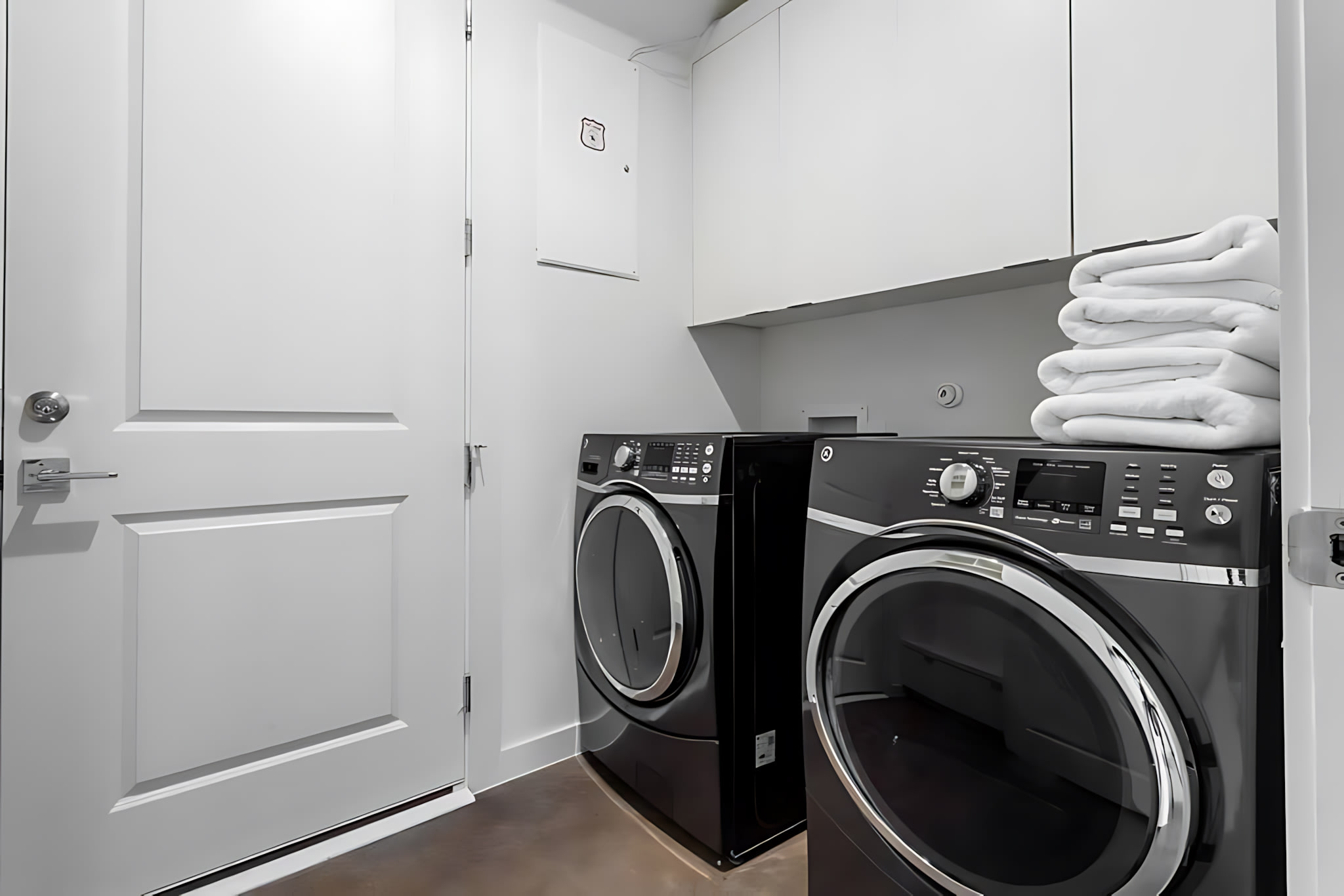 A modern laundry room featuring two sleek black front-loading washing machines and dryers, complemented by white upper cabinets and a stack of neatly folded white towels. A white paneled door is visible on the left, and the floor is a dark, smooth surface.