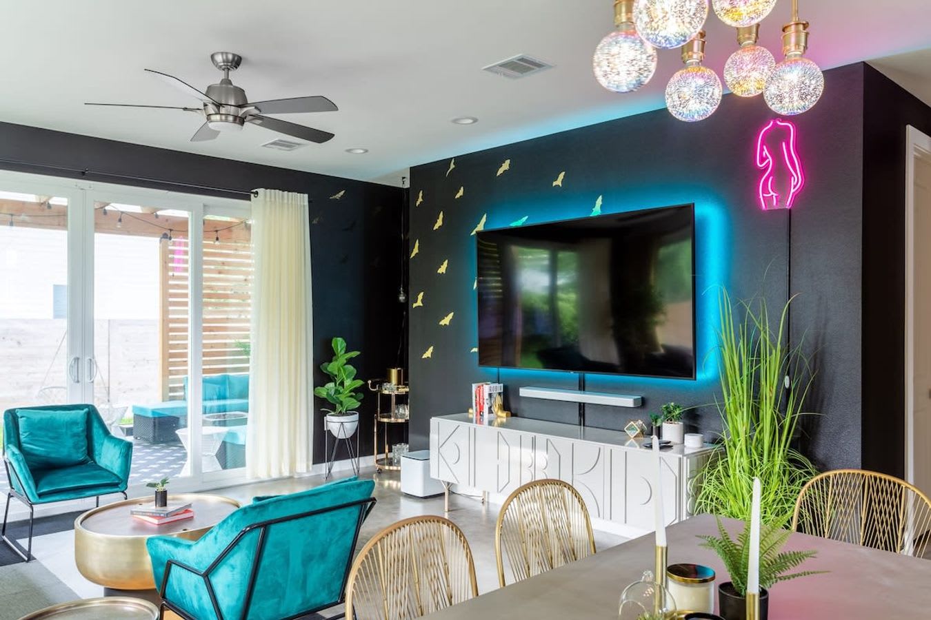 Vibrant living area with a large wall-mounted TV backed by blue neon lighting and golden bat decals on a black wall. Teal armchairs and a brass coffee table are visible, with a dining area and a unique chandelier in the foreground.