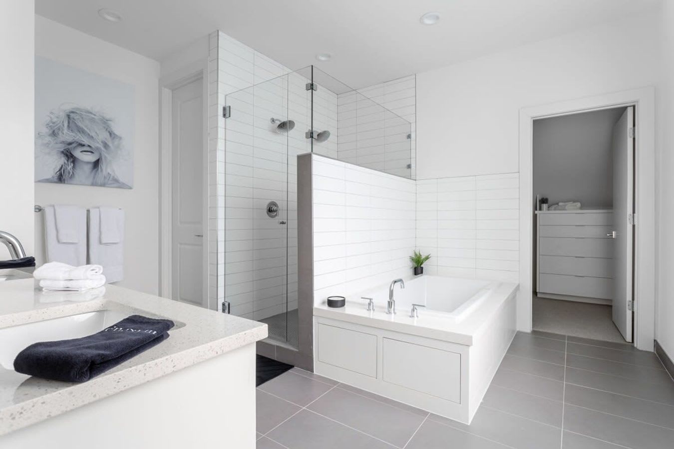 Full view of a bright, modern bathroom with a walk-in shower, a built-in bathtub, and a double vanity. White subway tiles and grey floor tiles create a clean aesthetic, with a large abstract artwork on the wall.