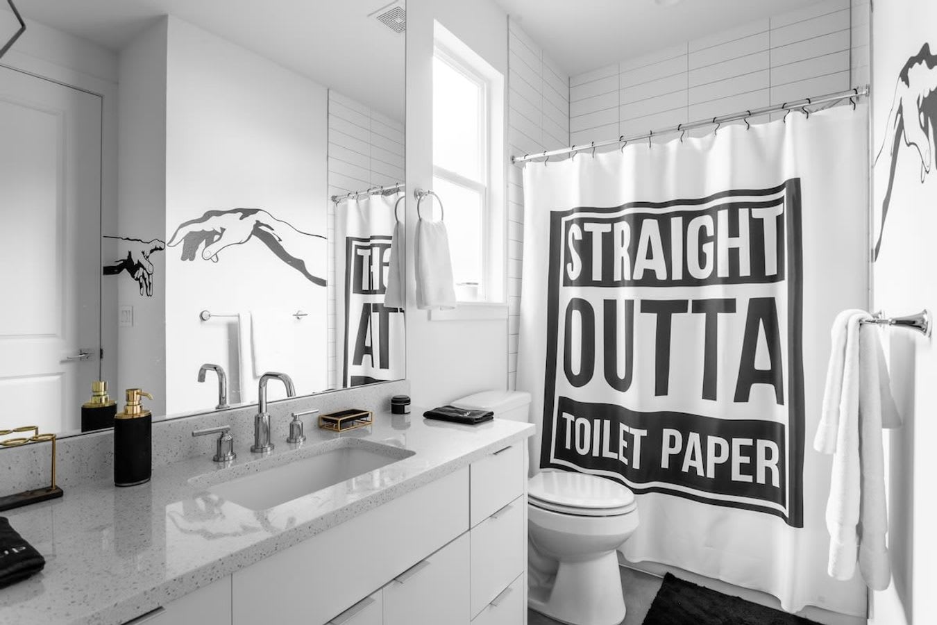 A bright white bathroom with a modern vanity, large mirror, and a shower featuring a "STRAIGHT OUTTA TOILET PAPER" shower curtain. Black graphic art adorns the wall above the toilet.