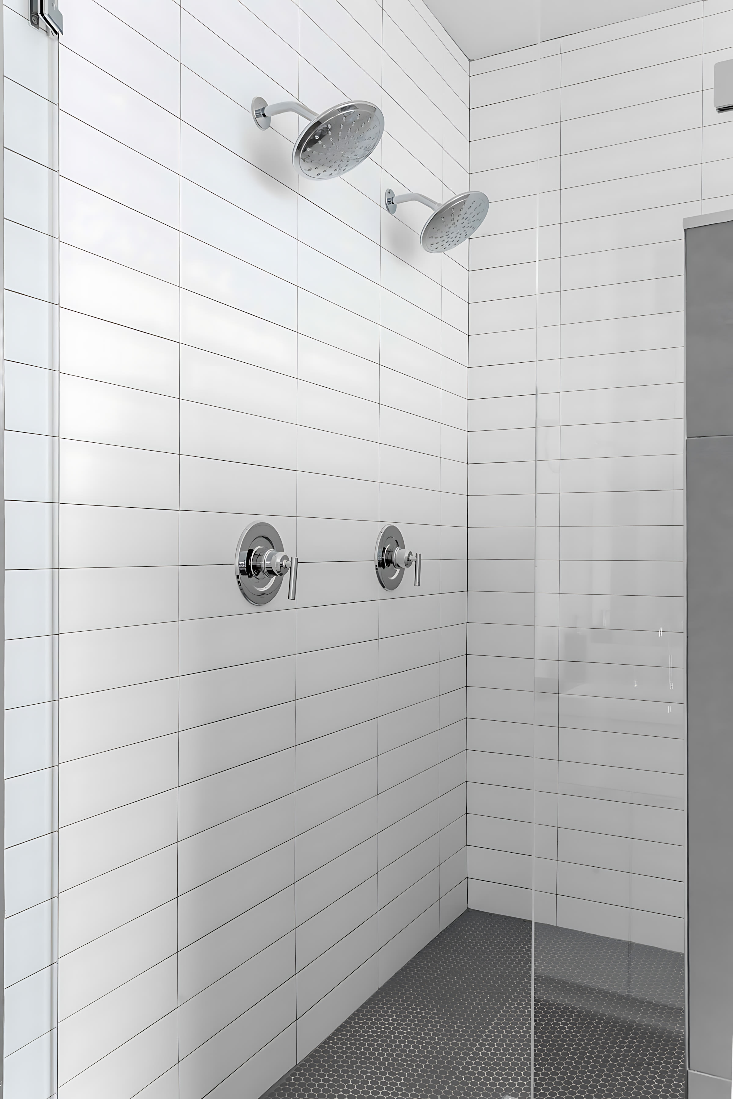 Spacious walk-in shower with white subway tiles on the walls and a grey penny tile floor. It features two rain shower heads and chrome fixtures, enclosed by a clear glass panel.