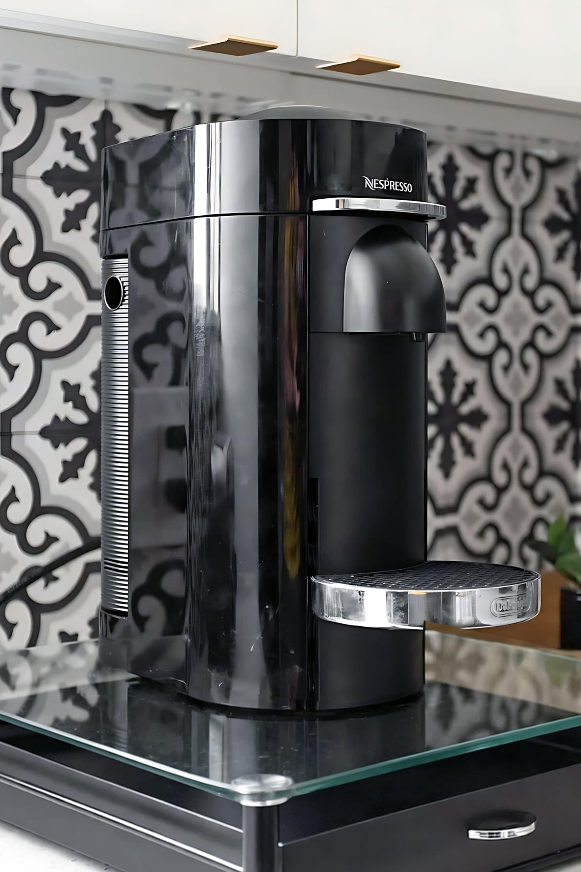 Close-up of a sleek black Nespresso coffee machine on a glass tray, set against a black and white patterned tile backsplash in a modern kitchen.
