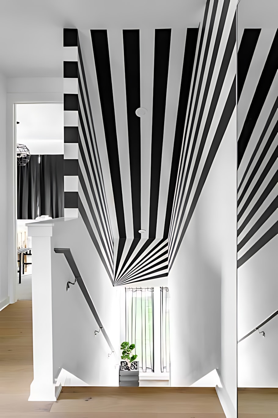 A dramatic staircase and hallway featuring bold black and white stripes extending from the wall onto the ceiling, creating an optical illusion. A small plant sits on the landing below.