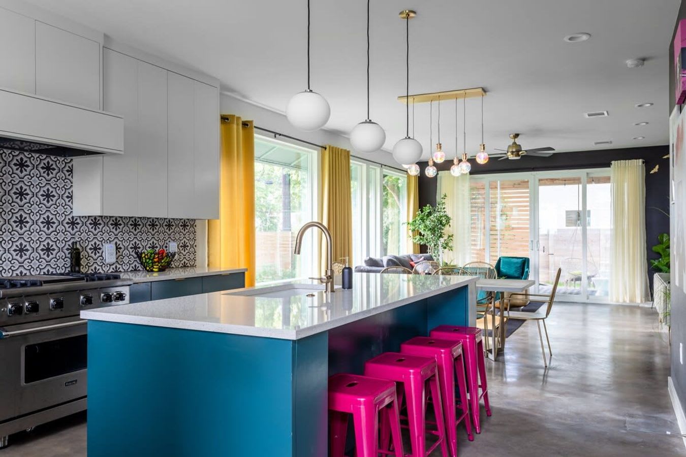 Modern kitchen with a large teal island, white quartz countertop, gold faucet, and four hot pink bar stools. The backsplash features black and white patterned tiles, and the space opens to a dining area and living room with yellow curtains.