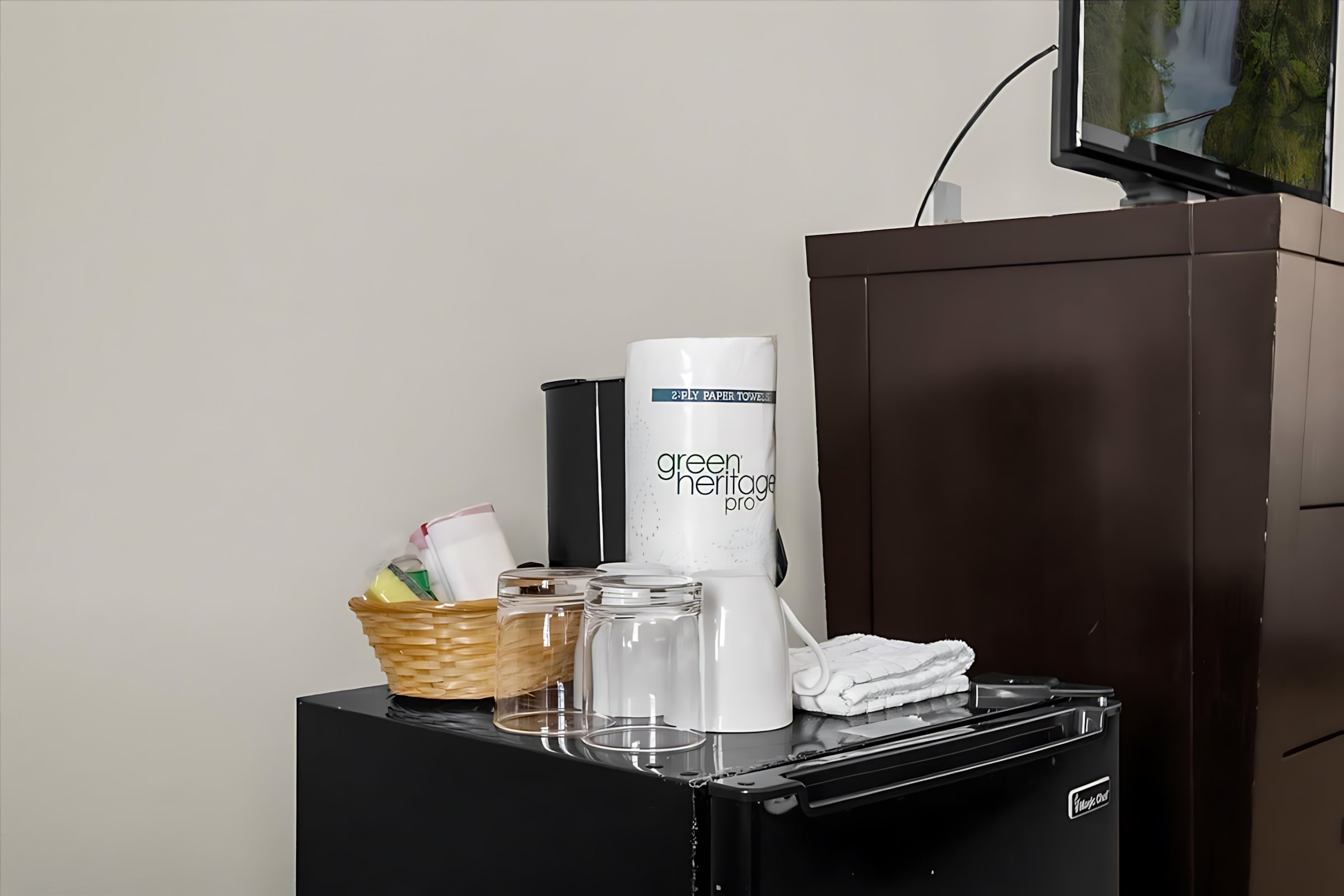 A black mini-fridge is topped with a roll of paper towels, clear drinking glasses, a white coffee maker, and a woven basket containing small items, set against a light wall.
