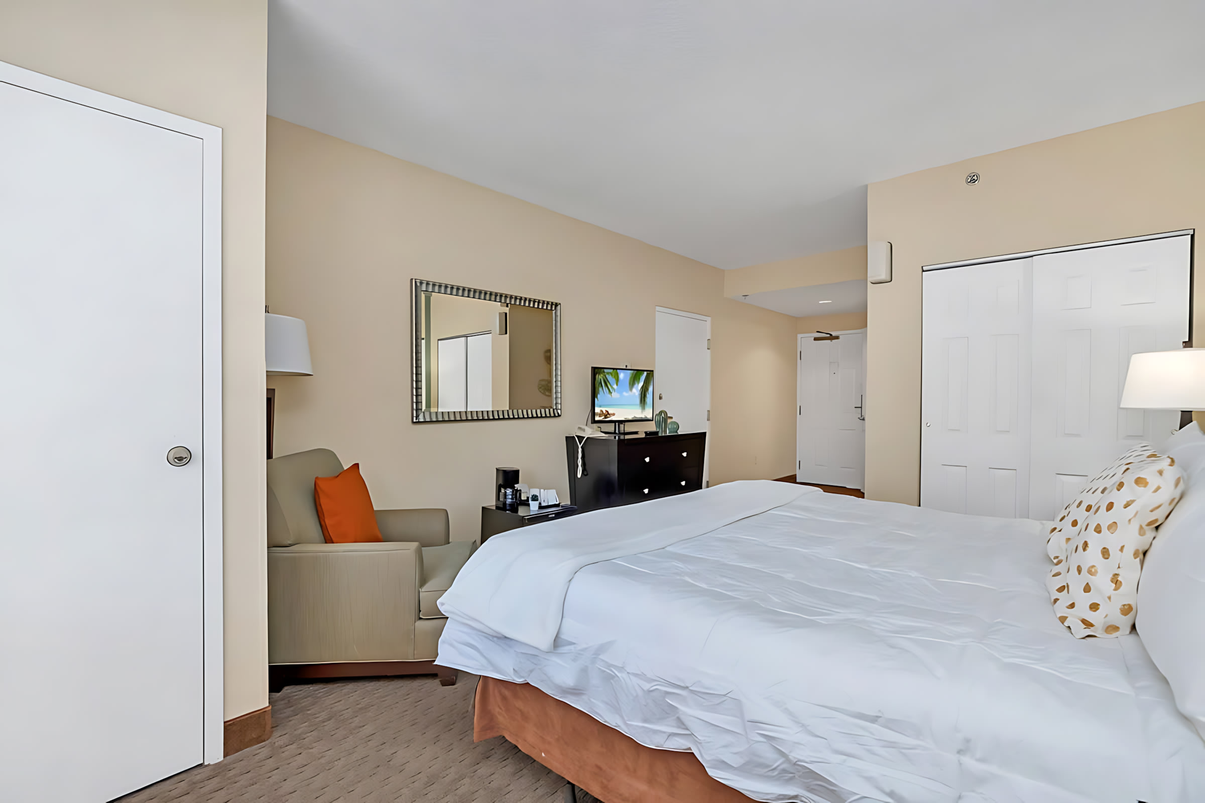 A king-sized bed with white bedding dominates the room, with an armchair and TV stand visible on the left, and a closet and entrance door on the right, all on a patterned carpet.