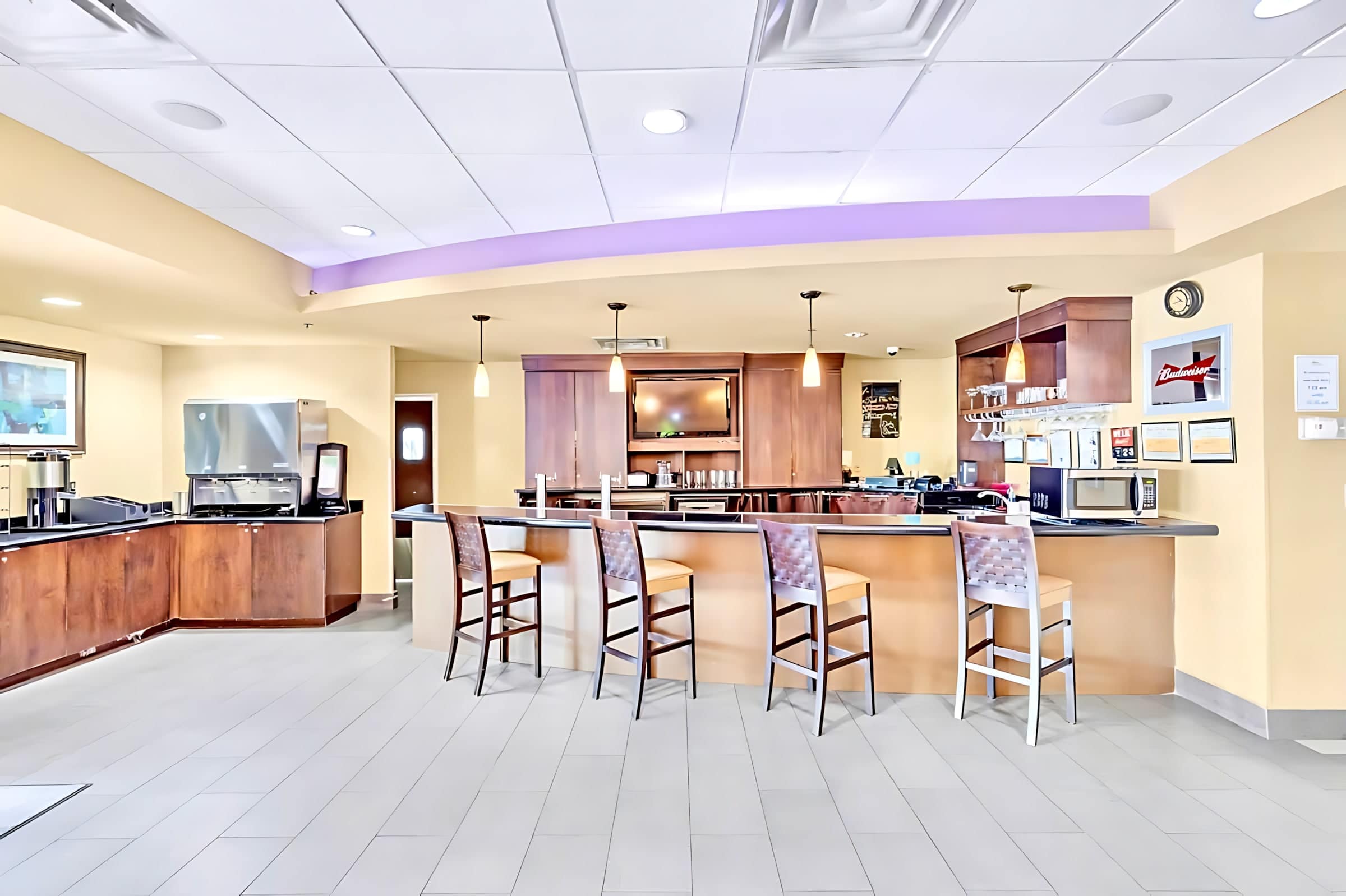 A modern communal bar and breakfast area featuring a long dark wood counter with several high-backed bar stools. Behind the counter are coffee machines, a microwave, and a television, all set against light beige walls.