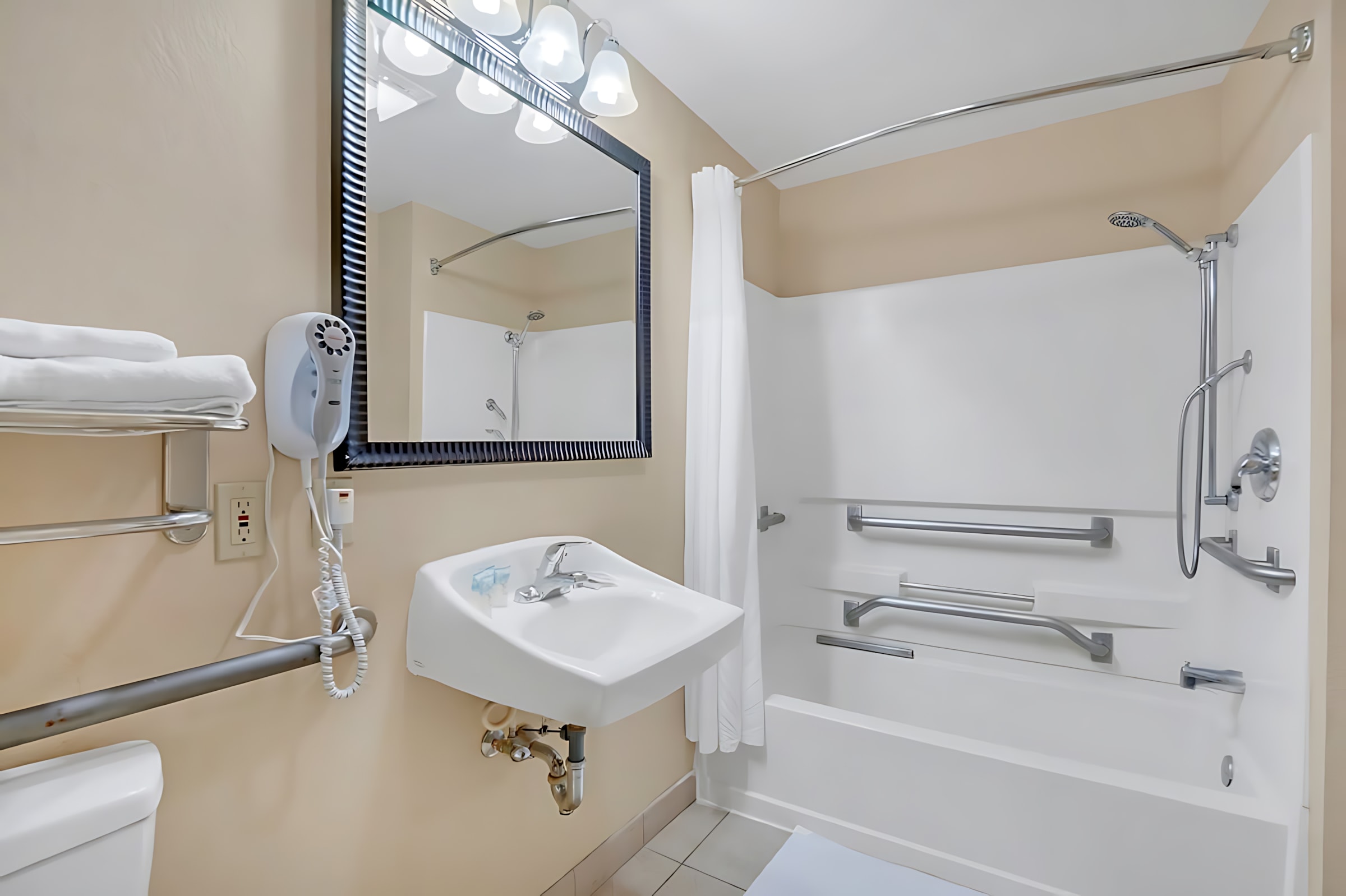 A clean bathroom with a white wall-mounted sink, toilet, and a bathtub with a shower head and grab bars, complemented by a large mirror and light-colored tiled floor.