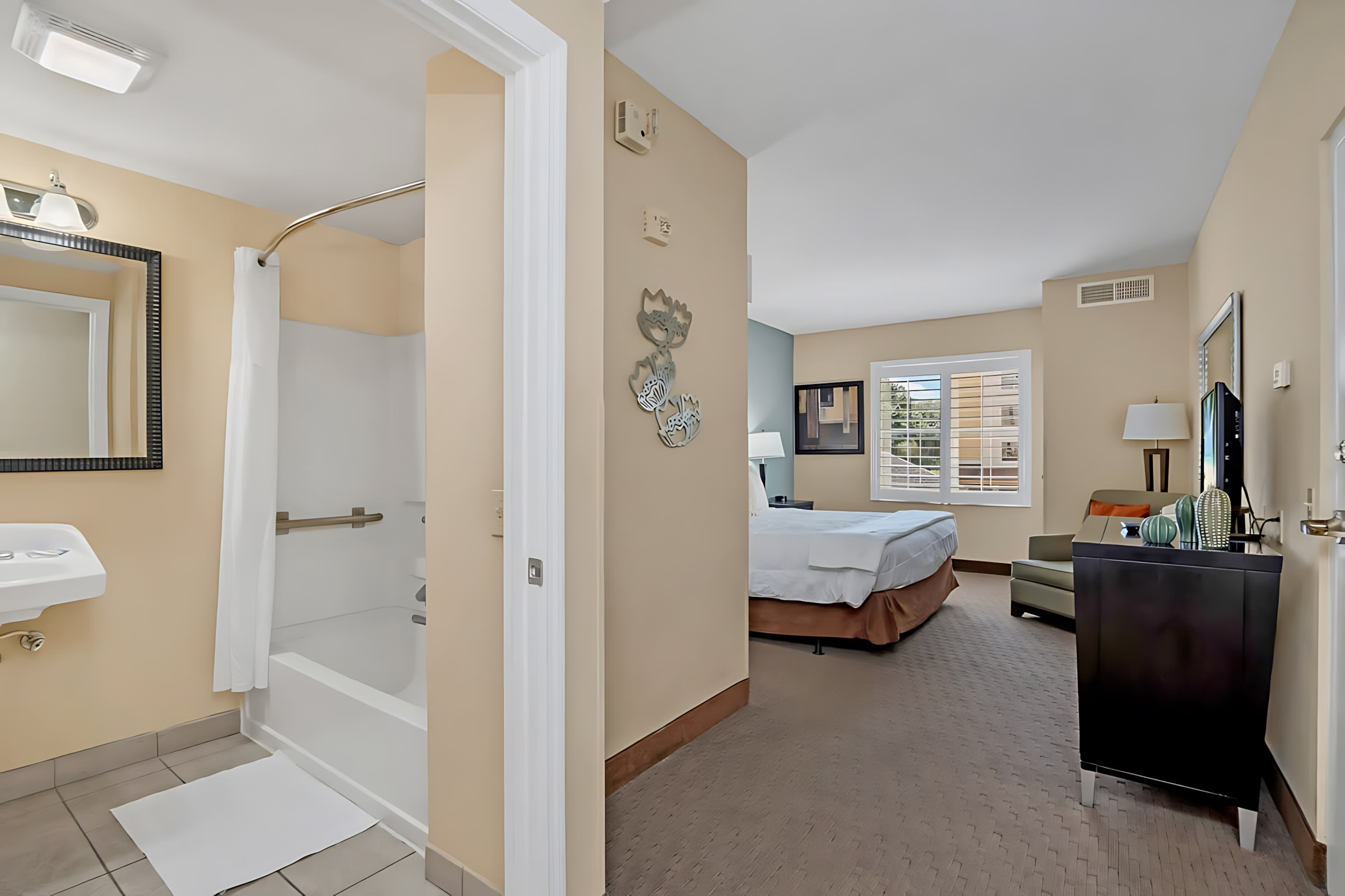 View from the bedroom into a bathroom with a white tub and shower curtain, while the bedroom features a king bed, armchair, and TV stand, all on a patterned carpet.