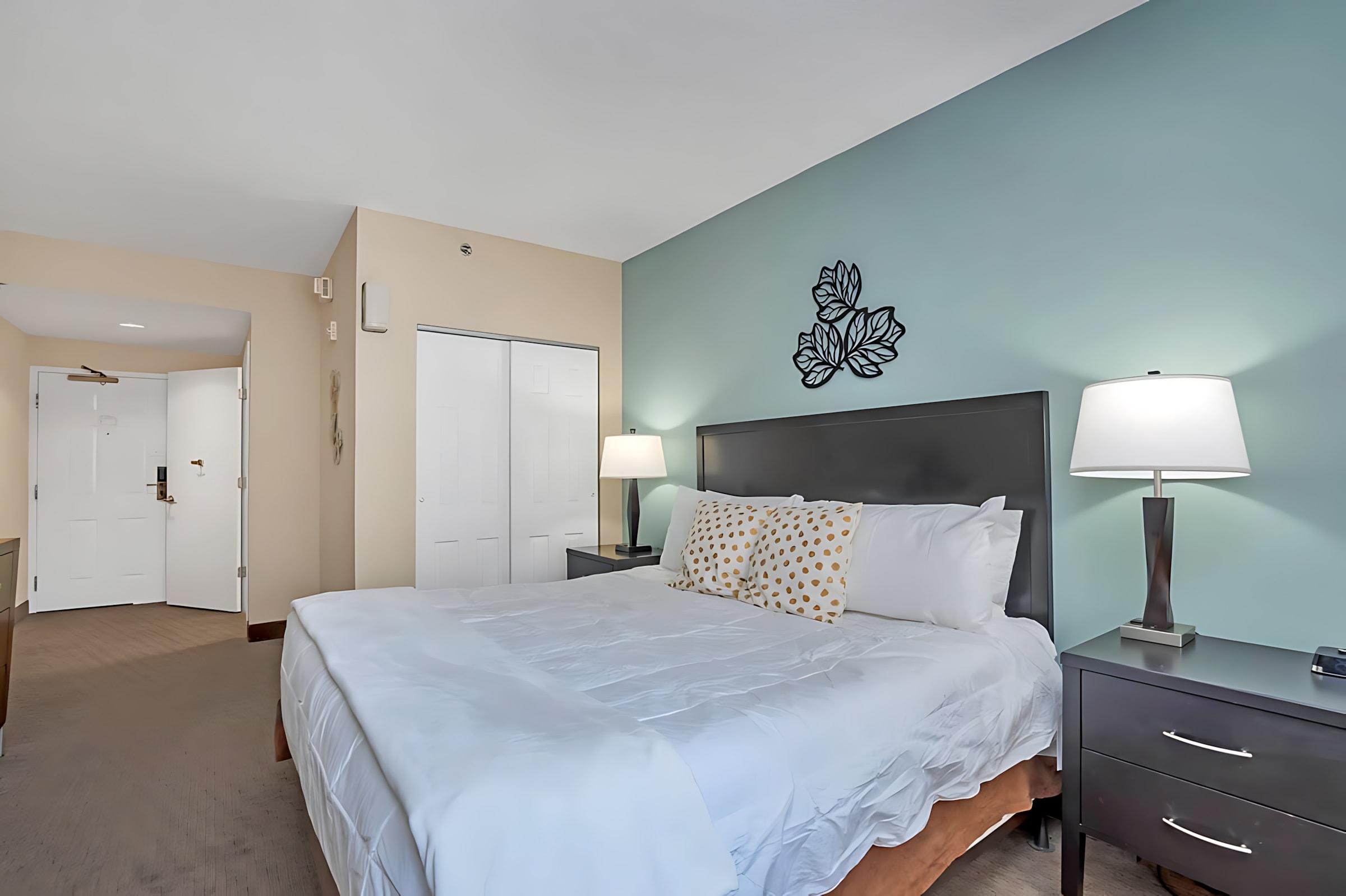 A spacious studio bedroom features a king-sized bed with white bedding against a teal accent wall, flanked by dark nightstands and lamps, with a closet and entrance door visible in the background.