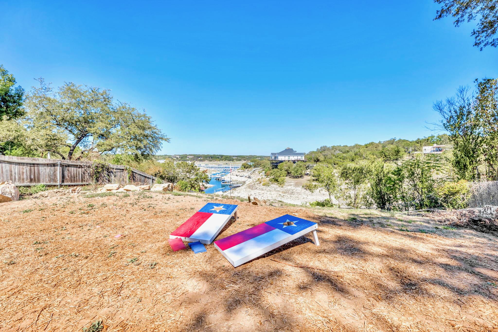 Two cornhole boards, painted with Texas flag designs, are set up on a dirt patch in the backyard. The background features a wide view of a lake and marina, with green trees and a clear blue sky.
