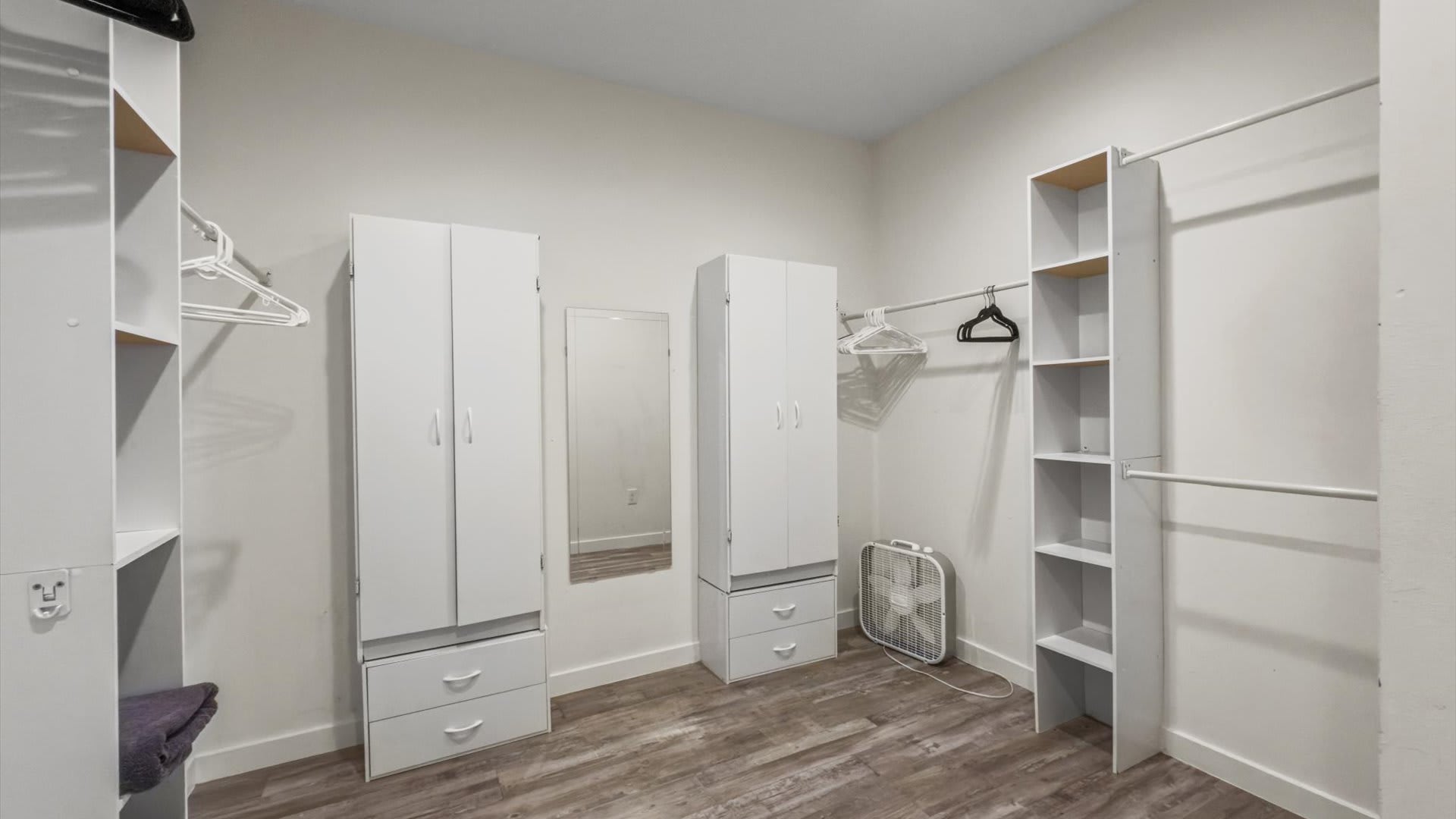 A spacious walk-in closet with white built-in shelving units, two white wardrobes with drawers, and ample hanging space for clothes. A full-length mirror and a floor fan are also present on the light wood-look flooring.