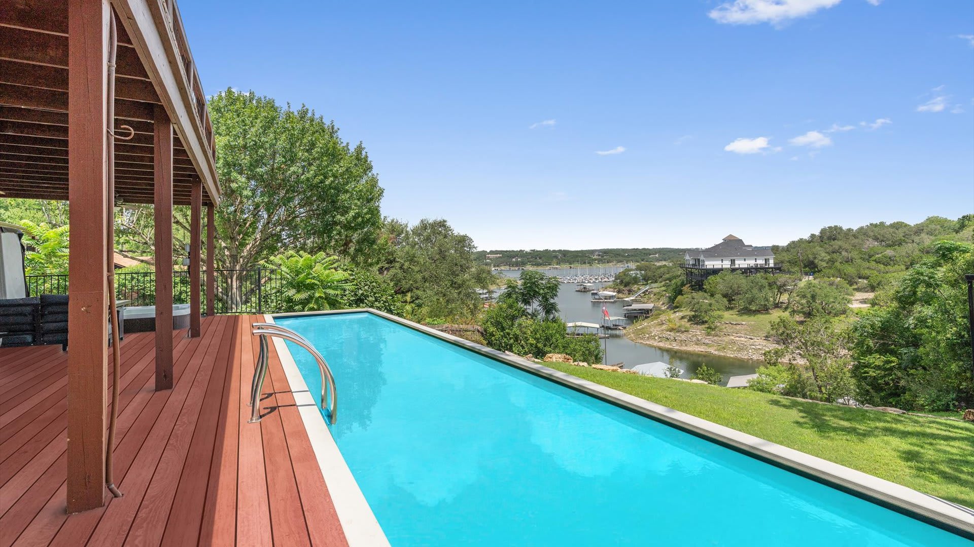 A long, narrow infinity-edge swimming pool with bright blue water is set into a wooden deck, offering panoramic views of a lake or river with boats and lush green hills under a clear blue sky.