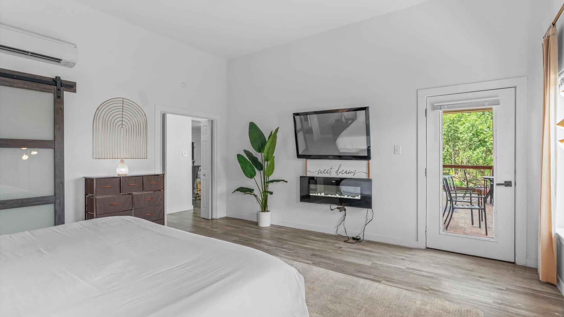 A modern bedroom with a large white bed, a wall-mounted TV above an electric fireplace, a tall green plant, and a glass door leading to an outdoor balcony with views of trees. The room features light hardwood floors and white walls.