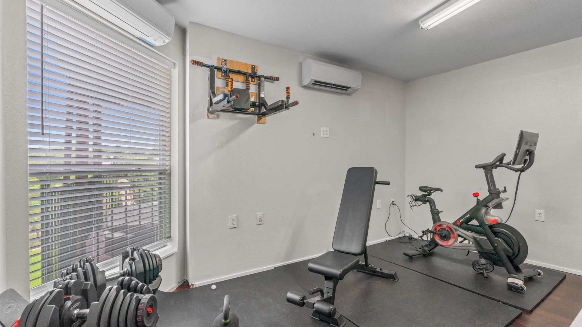 Home gym area with a Peloton bike, a weight bench, and various dumbbells on a black rubber mat. A window with blinds offers natural light, and a wall-mounted pull-up bar is visible.