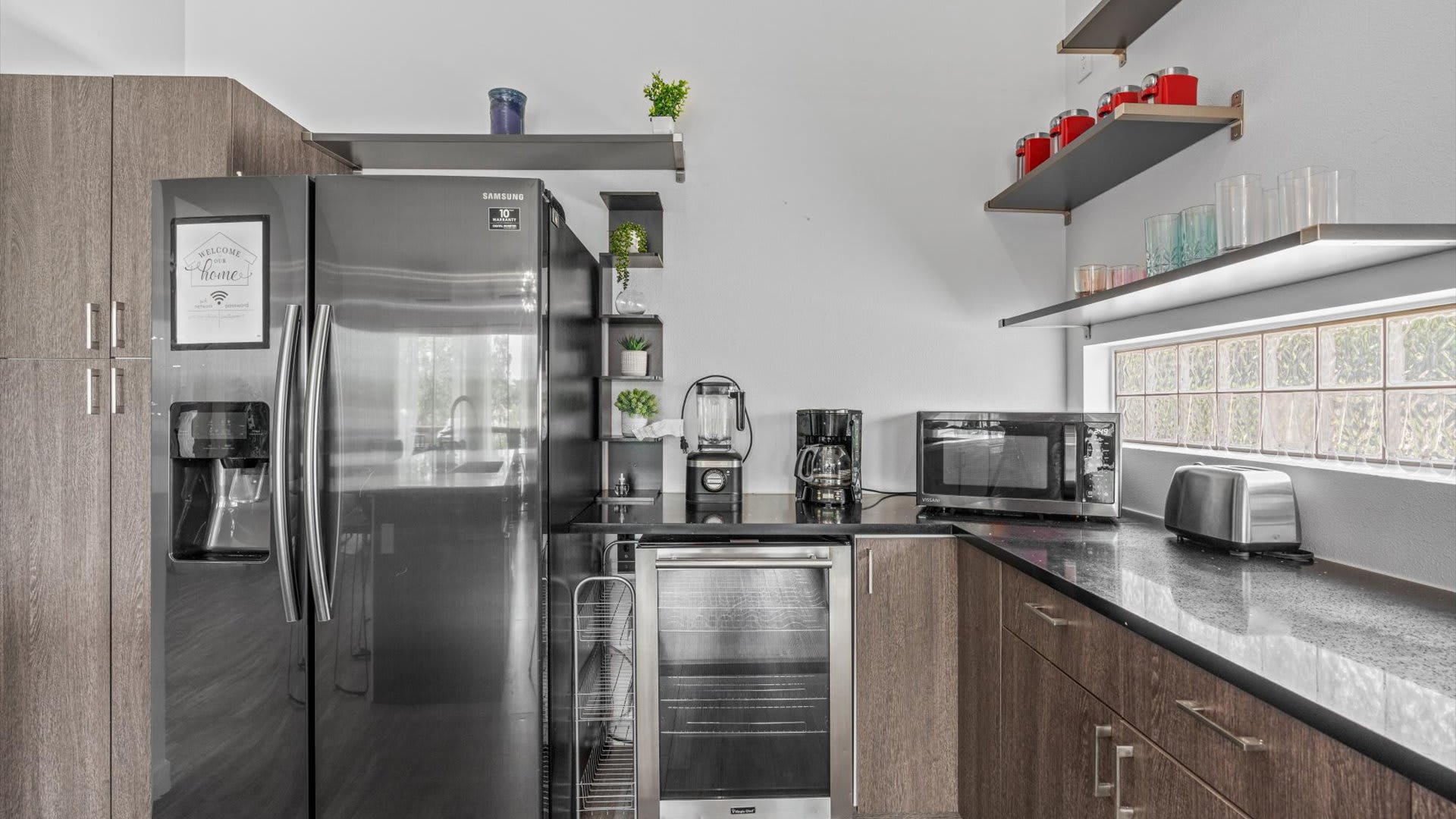 Another angle of the kitchen showing the stainless steel refrigerator, a beverage cooler, and various small appliances like a blender, coffee maker, and toaster on black countertops. Open shelves with decorative items are mounted on the white wall.