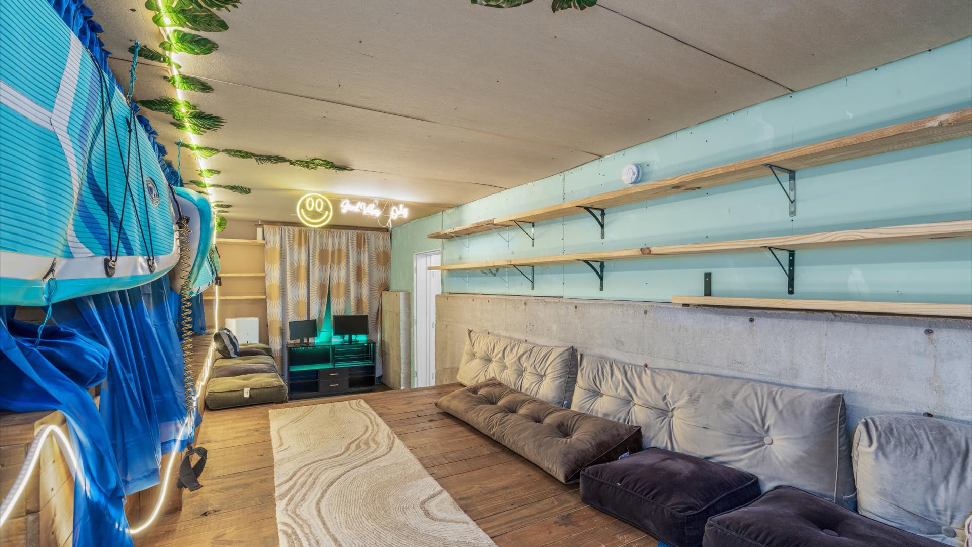 Another view of the recreation room shows a long row of floor cushions against a light blue wall with wooden shelves, opposite a section with paddleboards and blue curtains, all under a ceiling adorned with string lights and greenery.