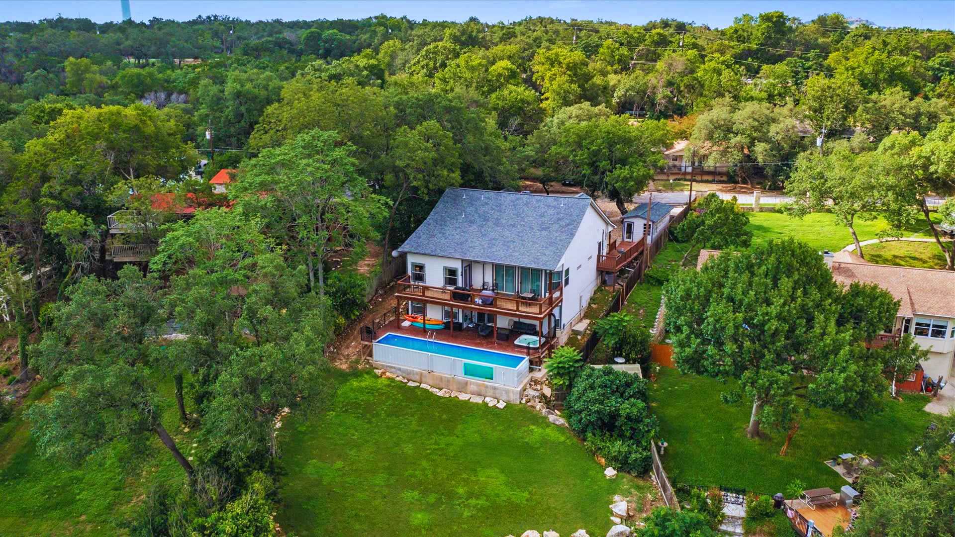 An aerial view captures a multi-story house with a light-colored exterior and dark roof, nestled among dense green trees, featuring a long swimming pool and a large wooden deck, all overlooking a scenic lake or river.