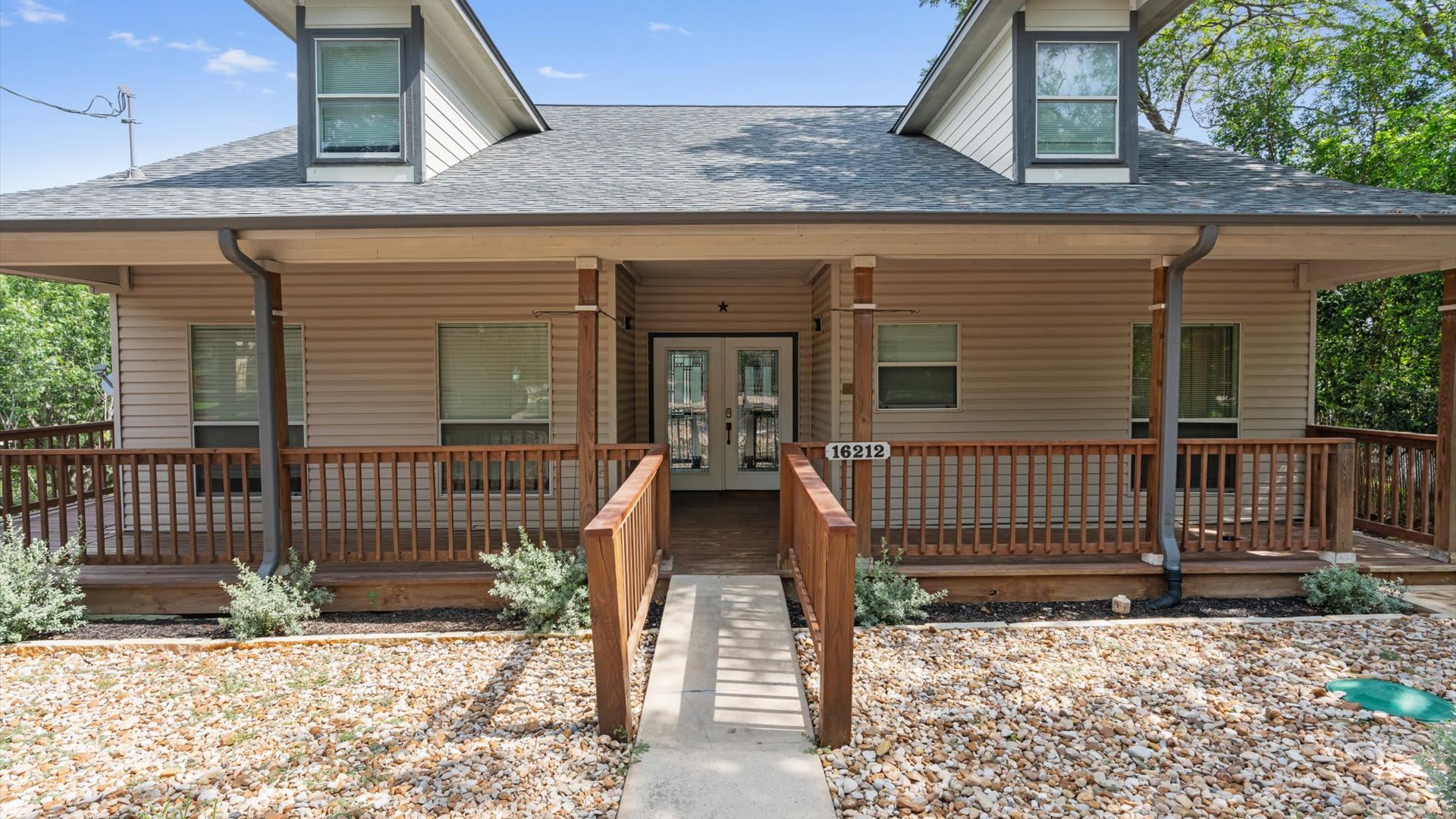 The front of a two-story house with light brown siding, a gray shingle roof, and a covered porch with wooden railings and columns. A concrete walkway leads to the double glass front doors, flanked by gravel landscaping.
