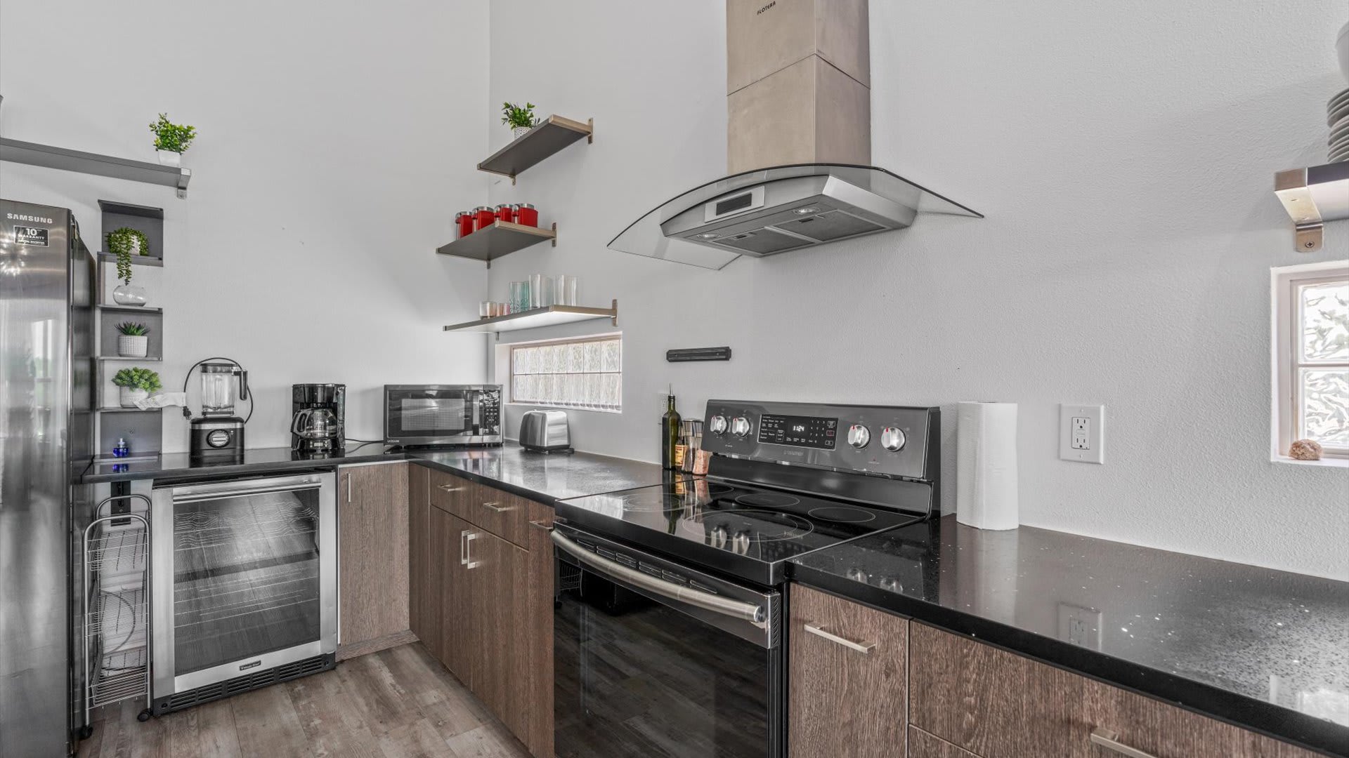 Detailed view of the kitchen counter with a black electric range, a microwave, toaster, and coffee maker. The area features black countertops, dark wood-look lower cabinets, and open shelving on white walls.